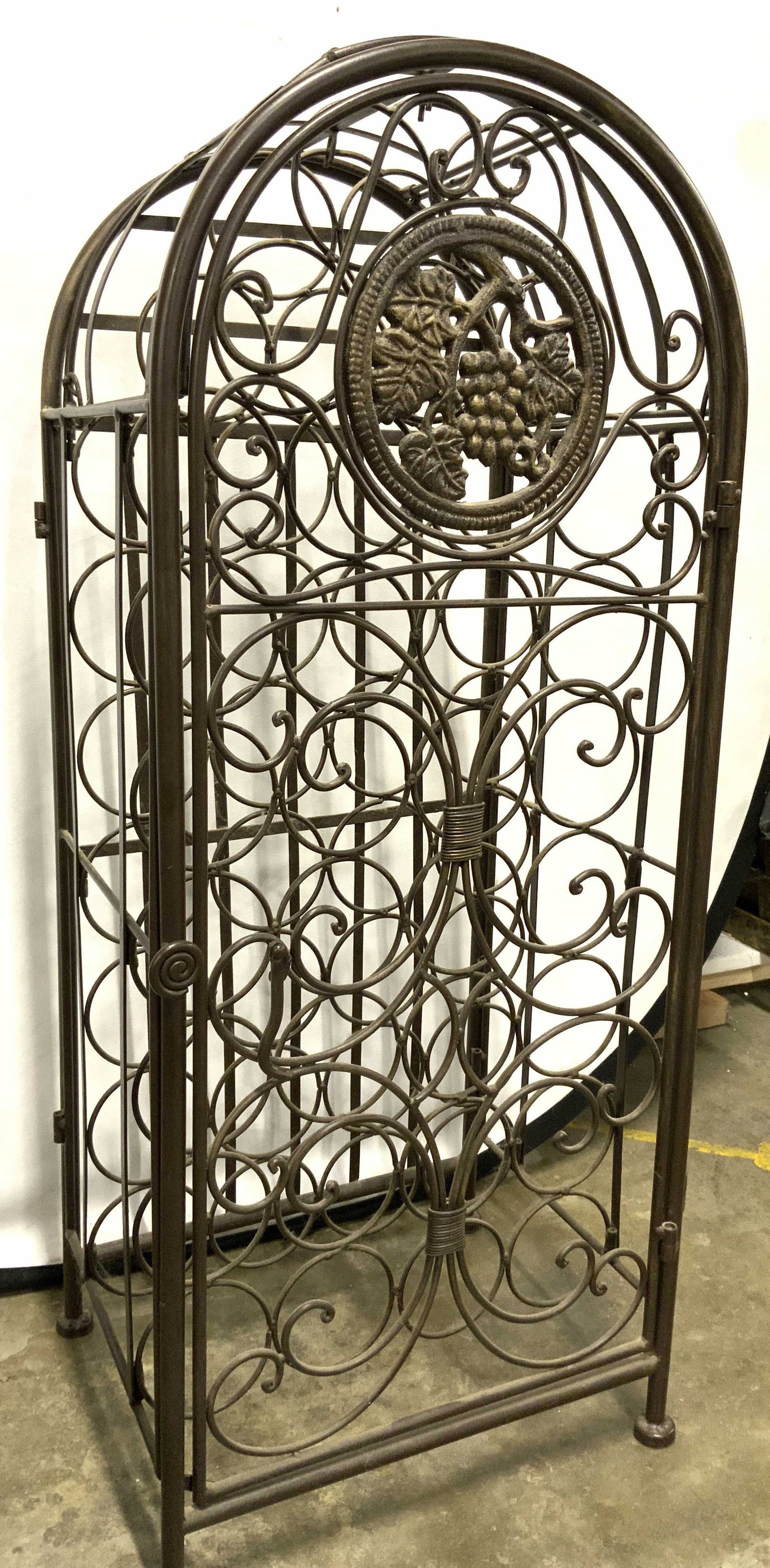 Arched Metal Wine Bottle Rack W Door: Bottle rack is brown toned and metal. Piece has arched form top. Front of piece has door. Top of piece is detachable. Door has grape, vine & leaf medallion detail. Measures approx 51 inches tall 17 in