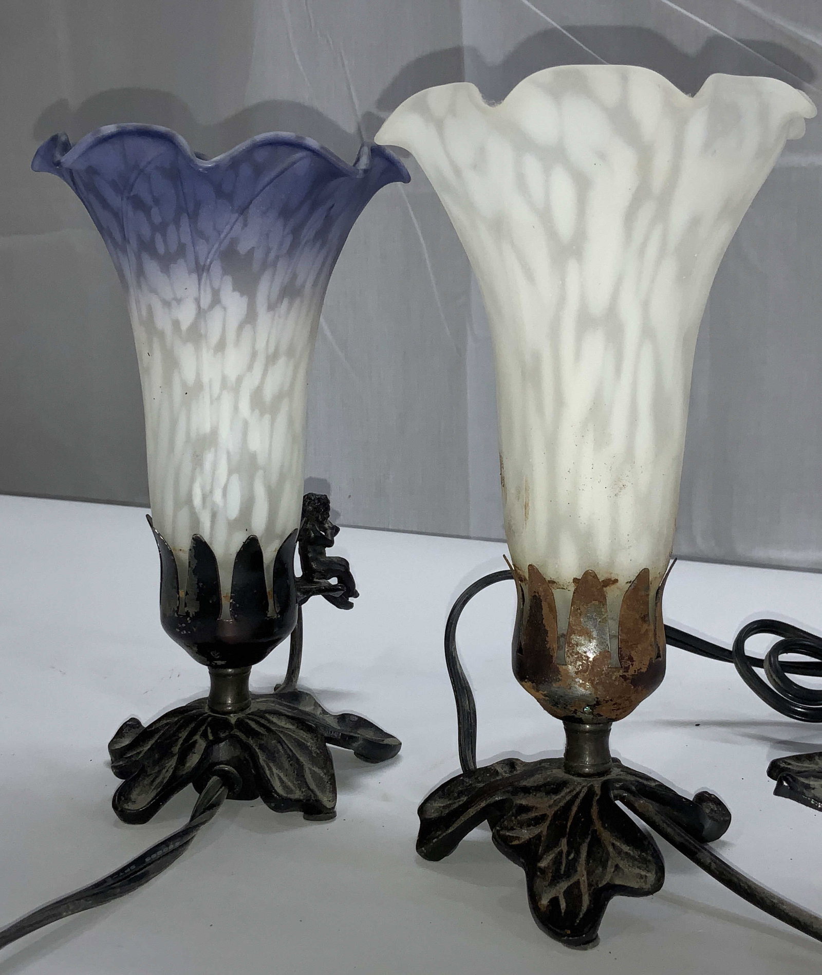 Near Pair Art Nouveau Frosted Art Glass Lights: Furled edge frosted art glass flower lamp shades with additional metal Figural to the side. Periwinkle and white shade has a cherub with wings playing the violin. All white art glass shade has a bird 