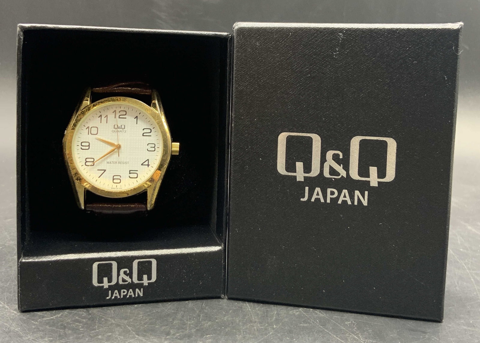 Q & Q JAPAN Brown Leather Wristwatch W/ Org Box: A brown leather, possibly leather style material, wristwatch. The watch face is white and framed by gold tone metal. Watch face reads Q & Q, QUARTZ. Comes in its original black box which reads Q & Q J