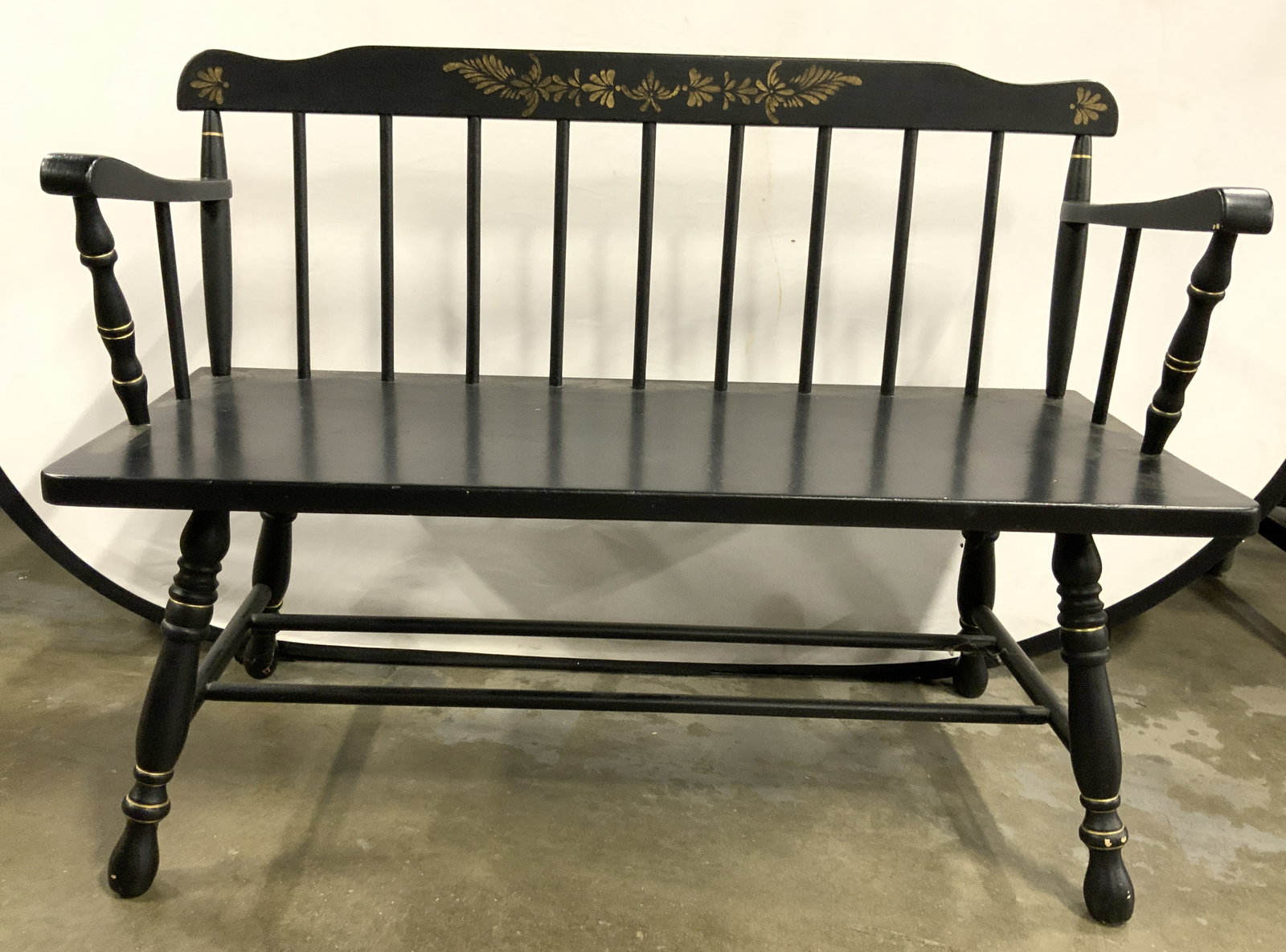 Vintage American Colonial Hitchcock Style Bench: Bench is vintage. American Colonial Hitchcock style bench. Bench is black toned and wooden. Piece has spindle backrest. Bench has armrests. Piece has 4 carved wooden legs with stretchers in between. B