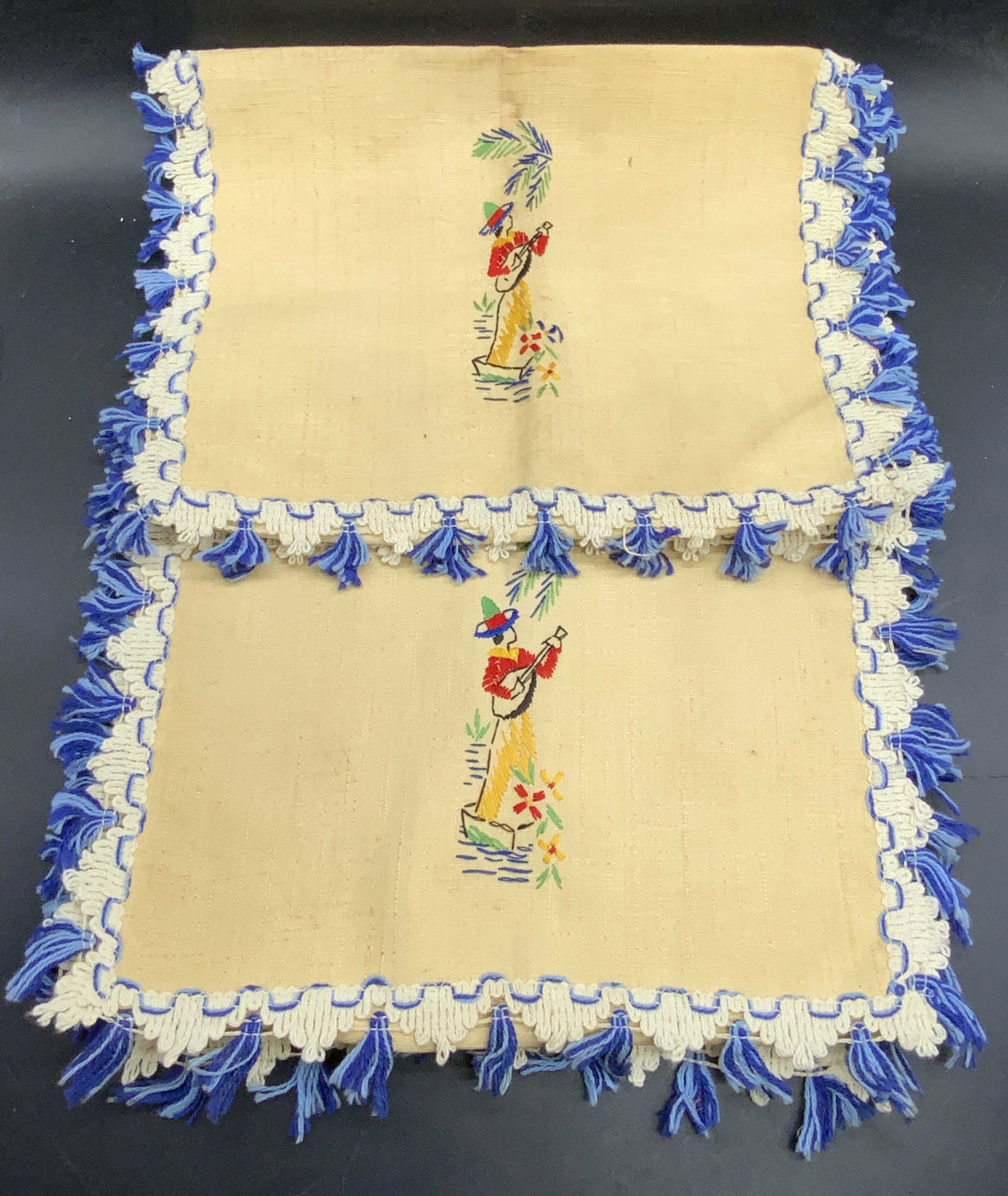 Pair Vintage Embroidered Table Linens: A pair of vintage embroidered table linens. Central motifs feature a person playing a musical instrument in a nature setting. Tassel trim. Measures approx. 43L x 15W in. Some surface wear and discolor