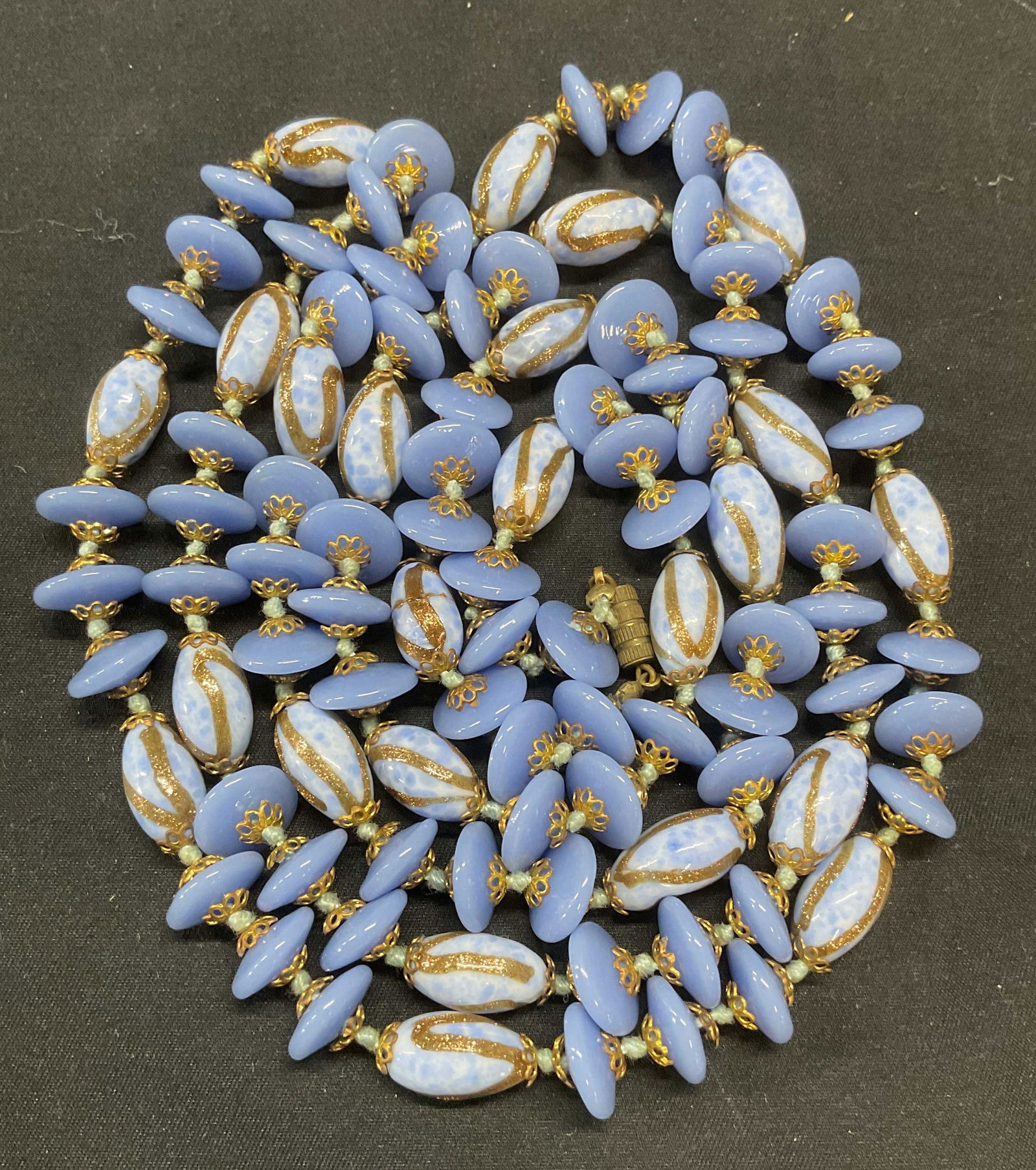 Vintage Blue Art Glass Beaded Necklace: Vintage blue periwinkle toned art glass beaded necklace with gold tone accents on double knotted string. Drop length approx. 25 inches. Some surface wear including scratching observed. Crack to one be