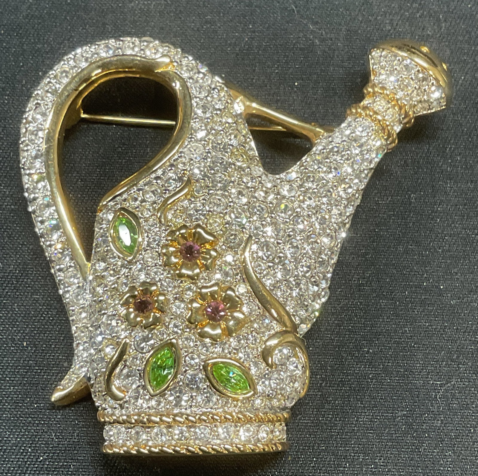 Swarovski Gold Pl Crystal Watering Can Brooch: Swarovski gold plated watering can motif brooch with floral design. Colorless, green, and purple toned crystals. Hinge pin to underside. Signed on side. Measures approx. 2 x 2.25 inches. Some surface 