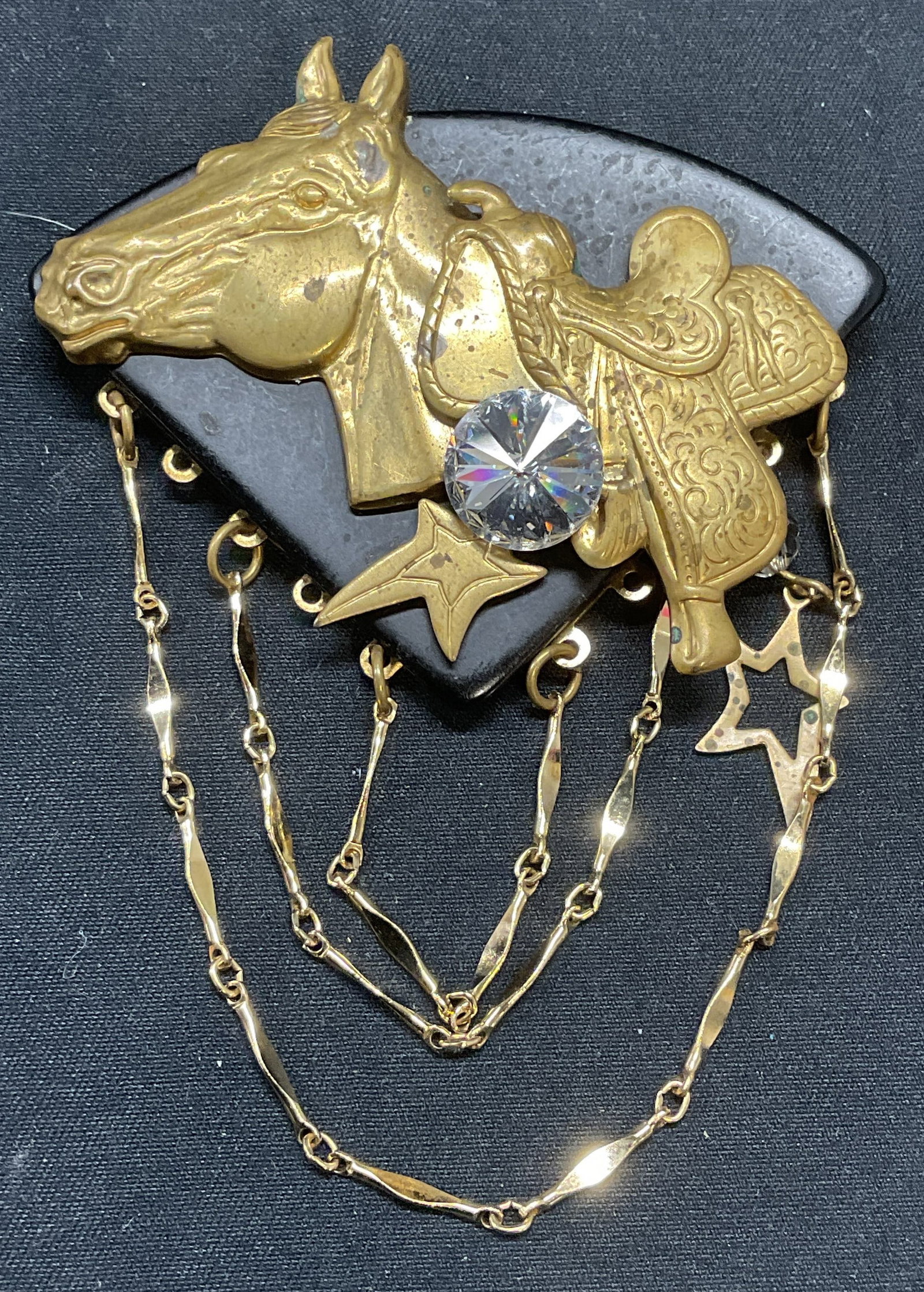 Gold Tone Crystal Horse Brooch, 3.75 Inches: Black composite and gold tone metal horse and saddle motif brooch with chain and colorless crystal decoration. Hinge pin to underside. Measures approx. 3.75 x 2.75 inches. Some surface wear including 