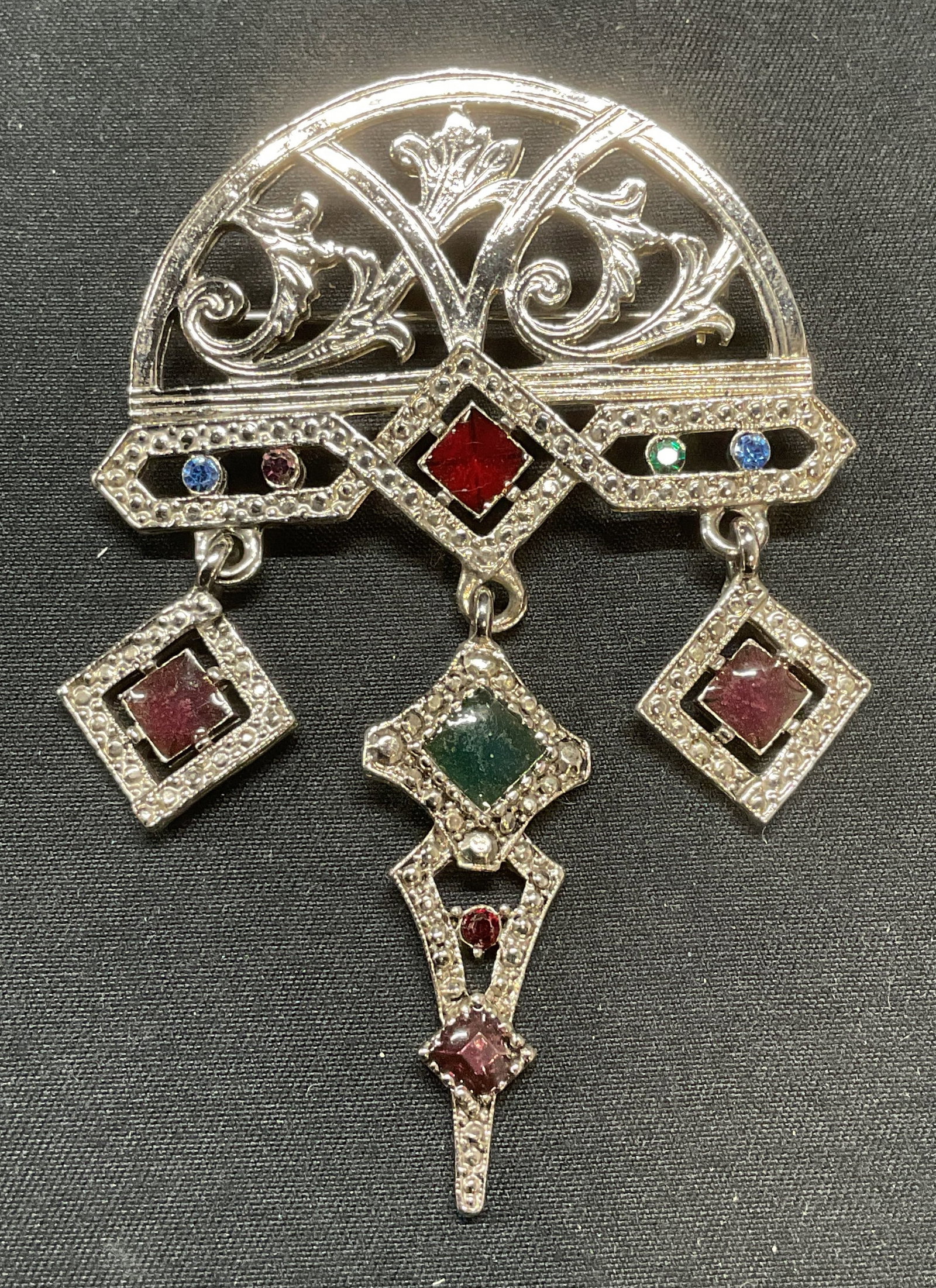 Silver Tone Art Deco Style Crystal Brooch, 3.25In: Silver tone art deco style scroll motif chandelier brooch. Crystal and lacquer decoration in hues of blue, red, and green. Hinge pin to underside. Measures approx. 3.25 x 2.25 inches. Some surface wea