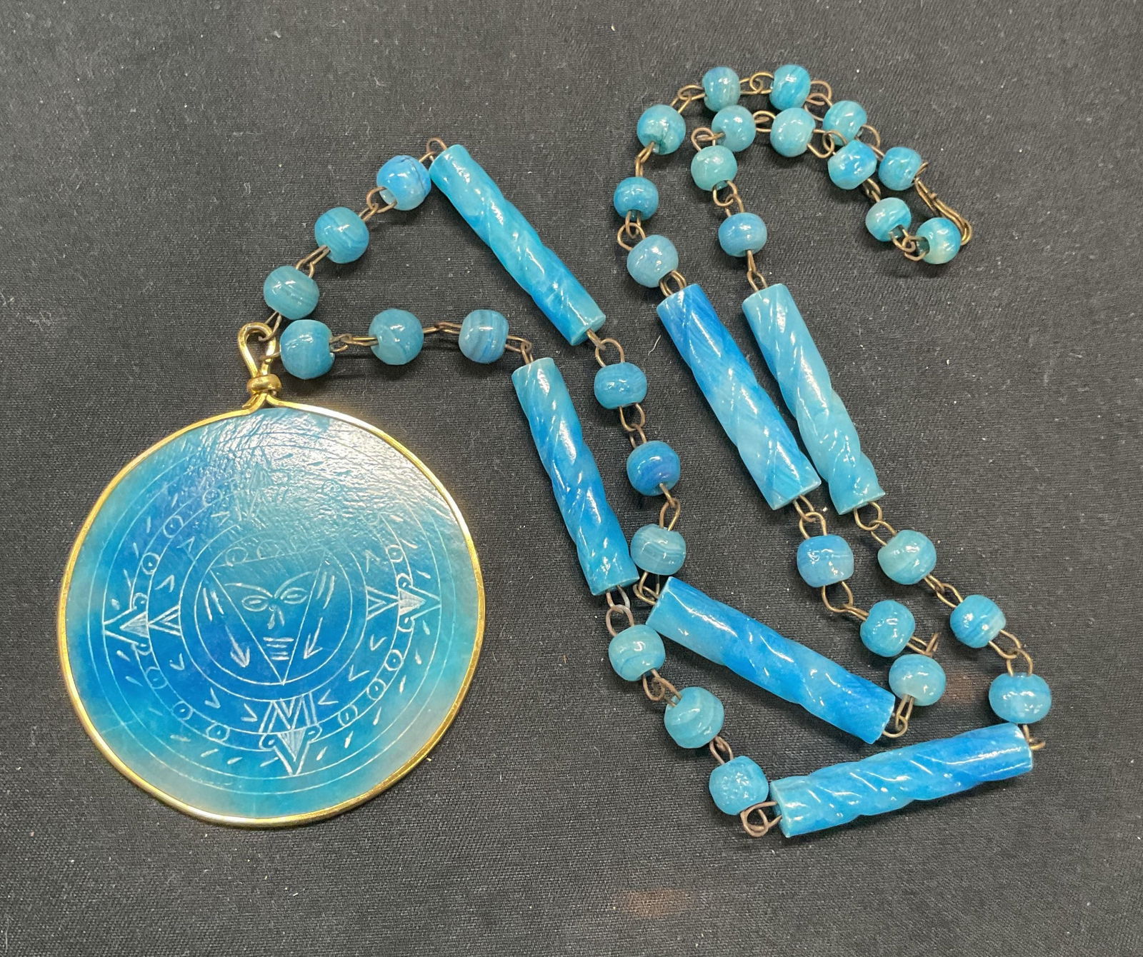 Etched Blue Stone Pendant Necklace: Blue stone beaded pendant necklace with etched tribal motif. Drop length approx. 12 inches. Pendant measures approx. 2 inches diameter. Some surface wear including scratching observed. Jewelry, estate