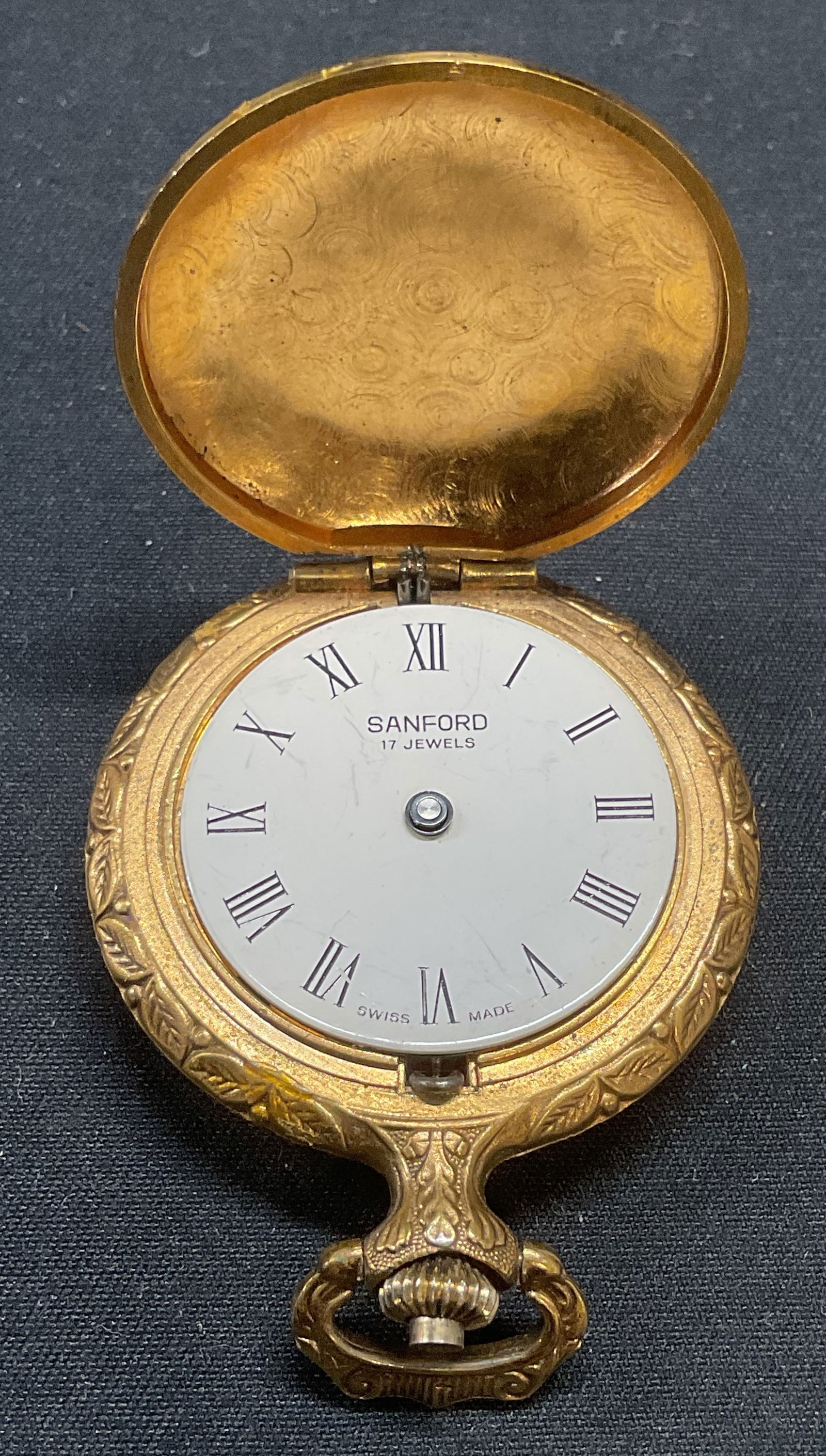 Sanford Gold Tone Figural & Clover Pocket Watch: Sanford gold tone pocket watch with raised figural, bird, and clover motif on case. Signed in watch face. Measures approx. 1.5 inches diameter. Not tested for function. Hand need replacement. Some sur