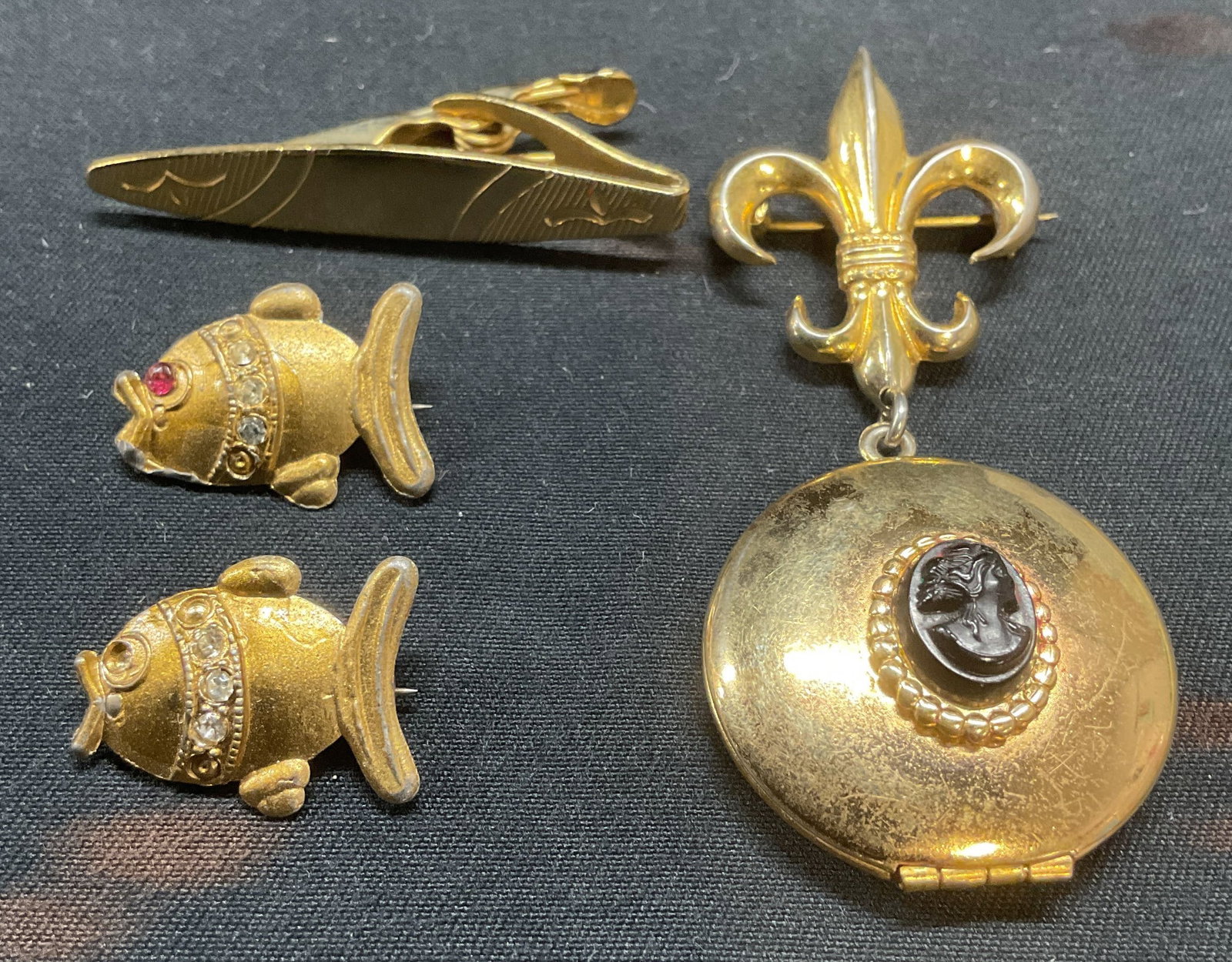 Lot 4 Fleur De Lis locket Brooch, Fish Brooches +: Lot of 4 jewelry pieces. Gold tone fleur de lis brooch with locket pendant and cameo decoration, 2 gold tone crystal fish motif brooches, and gold tone tie clip. Largest piece measures approx. 2.5 x 1