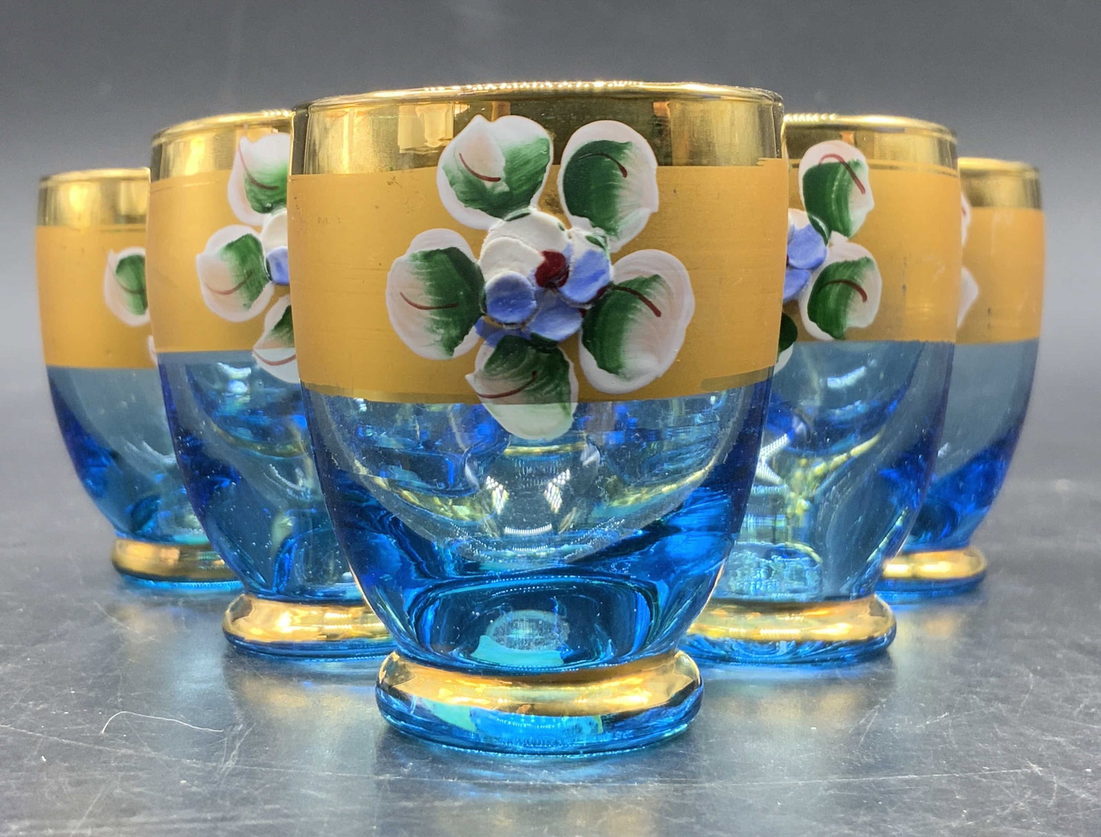 Set 6 Italian Hand Painted Art Glass Shot Glasses: A set of six hand painted floral motif blue shot glasses. The florals are raised. Gilt accents. Stickers to undersides read MADE IN ITALY. Measures approx. 2H x 1.75W in. Slight differences in sizes. 