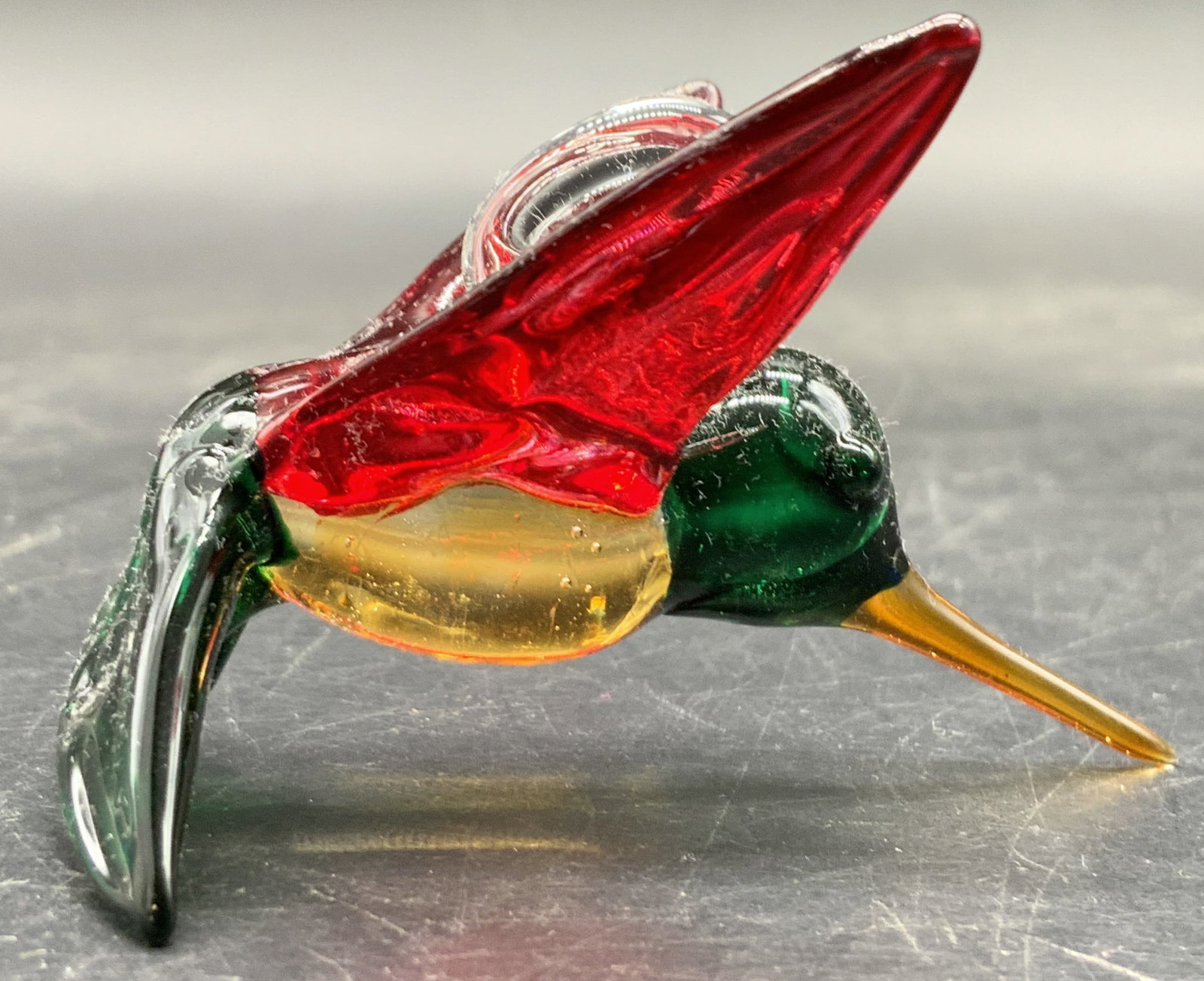 Art Glass Hummingbird Figural Ornament / Pendant: Art glass hummingbird figural ornament / pendant. Bail between the wings. Green, red, clear, and yellow hued glass to the piece. Measures approx. 2L x 2W in. Bubbles to the glass. Some surface wear co