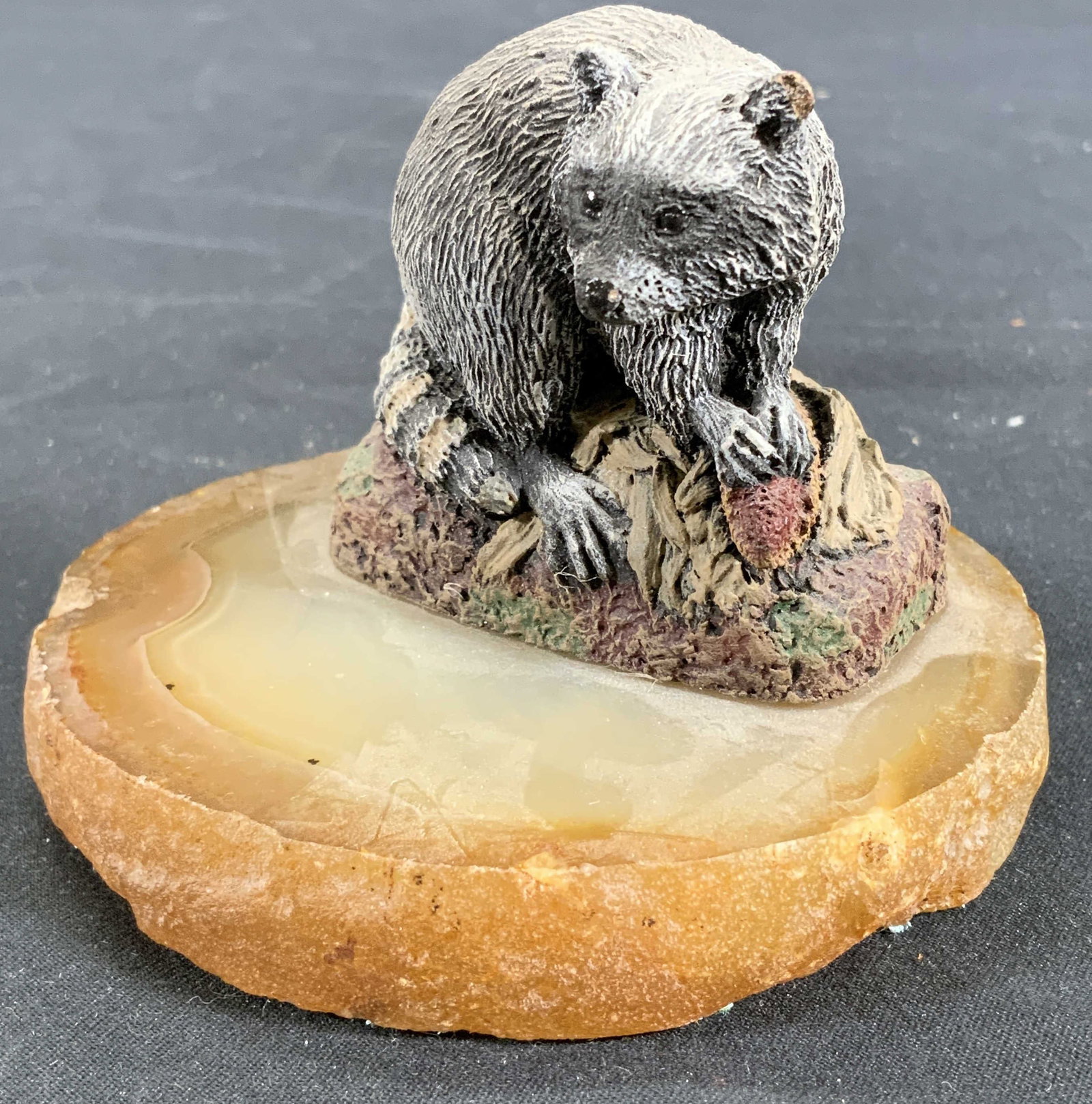 Miniature Raccoon Stealing Corn Figural on Geode: Composite miniature figural. Base is warm toned geode with felt base, miniature depicts a raccoon with a corn husk in its hands. Verso of figural has engraved inscription. Inscription illegible except