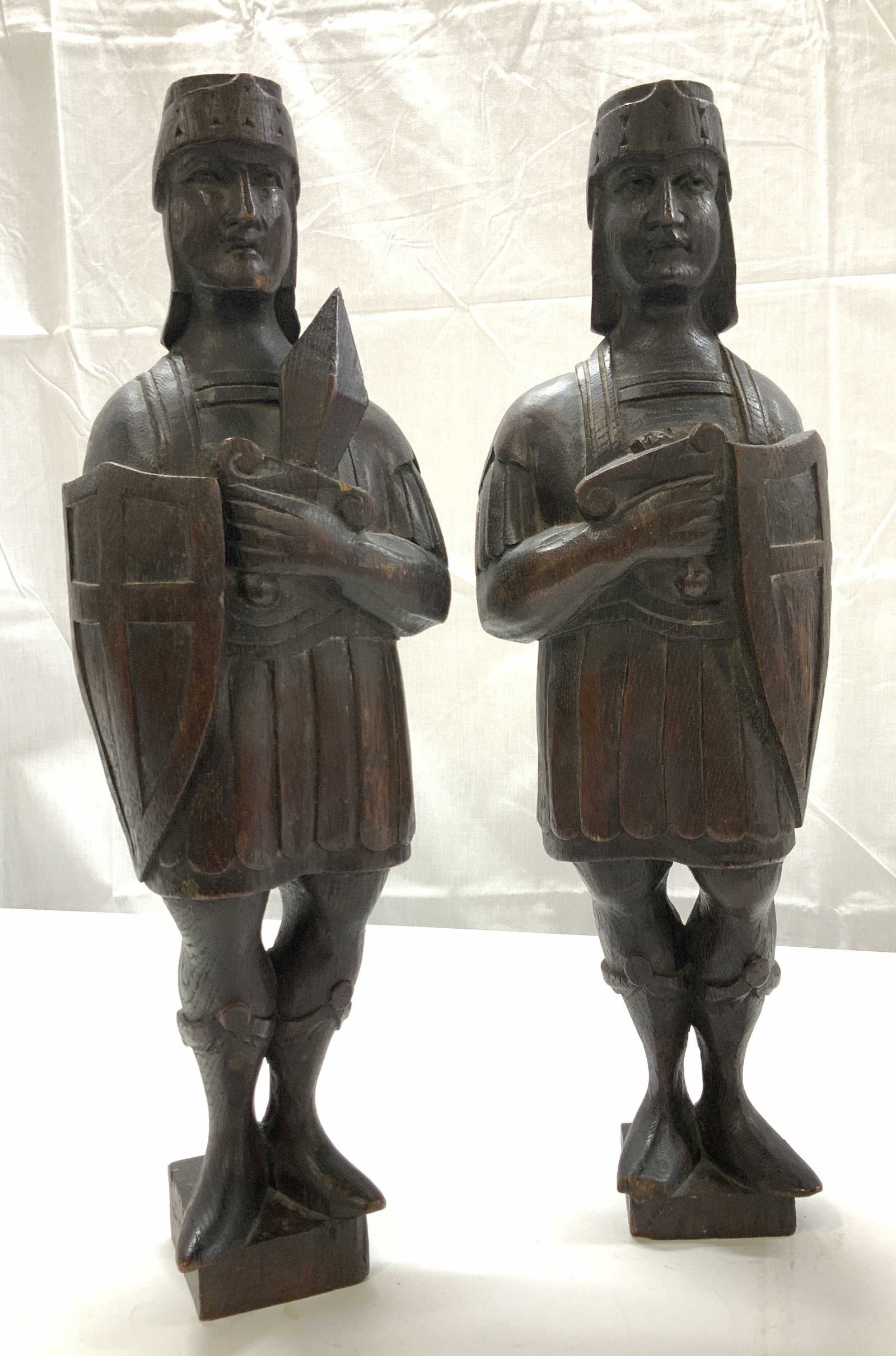 Pair Wooden Soldier Sculptures: Hand crafted vintage wooden soldiers wearing helmets, and battle uniforms with shields and sword. 18 in. Tall x 5.75 in. Wide. One soldier has a chipped foot and sword. Carved from one piece of wood. 