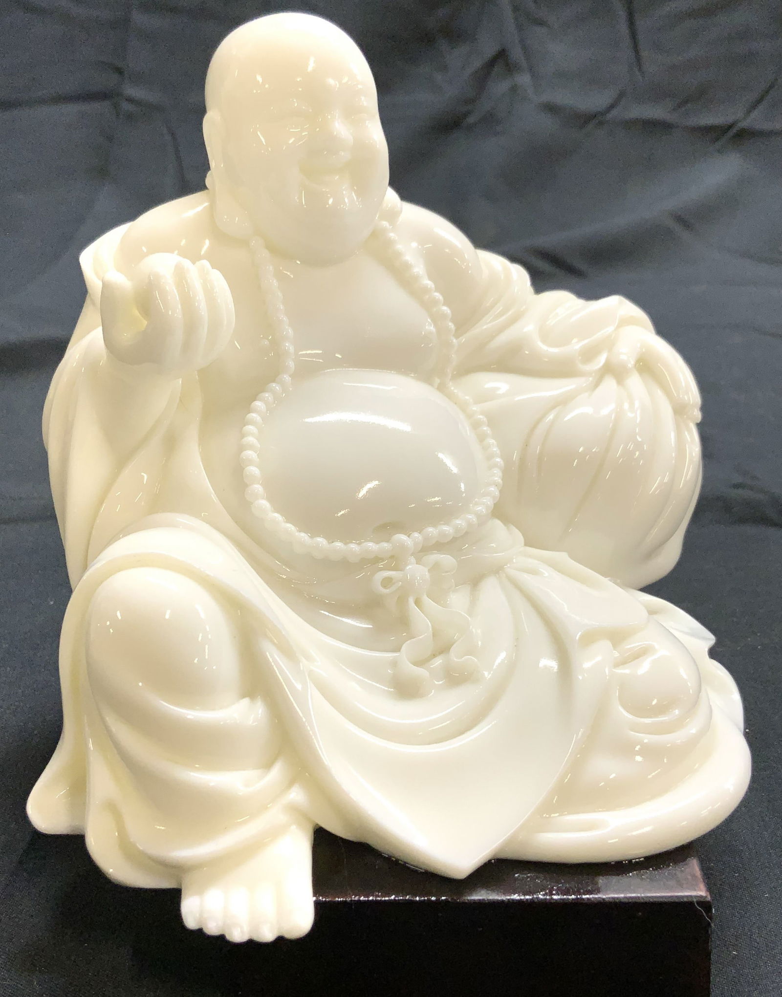 Milk Glass Buddha On Wood Base: Buddha figure is white toned, appears to be glass. Depicts a seated Buddha figure wearing beaded necklace & object in hand. Buddha figure is affixed to mahogany toned wooden base. Piece measures appro
