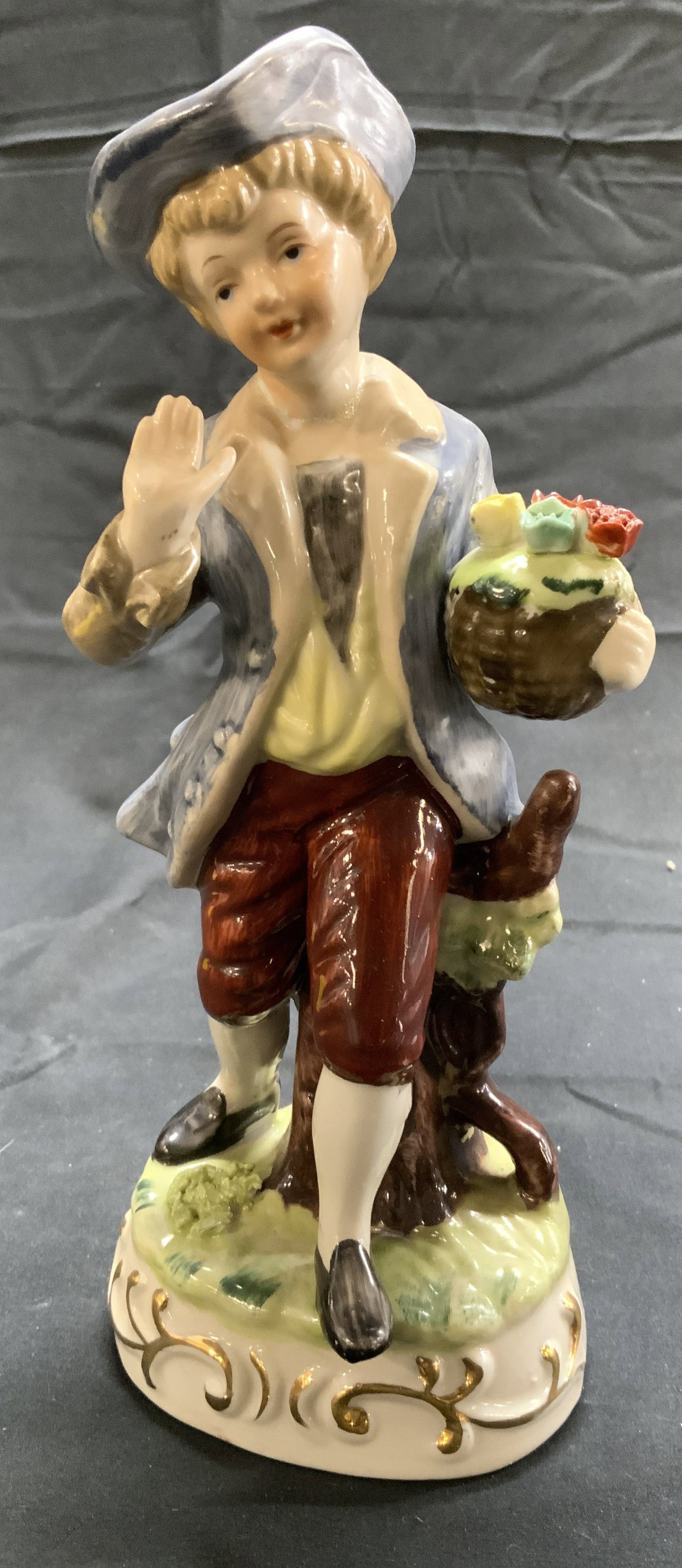 TOMA Vntg Porc Boy W Flowers Figural: Tag on Underside reads TOMA. Piece depicts a male figure holding a basket of flowers. Piece appears to possibly be hand painted. Base has gilt detail. Piece measures approximately 8.25 inches tall 3.2