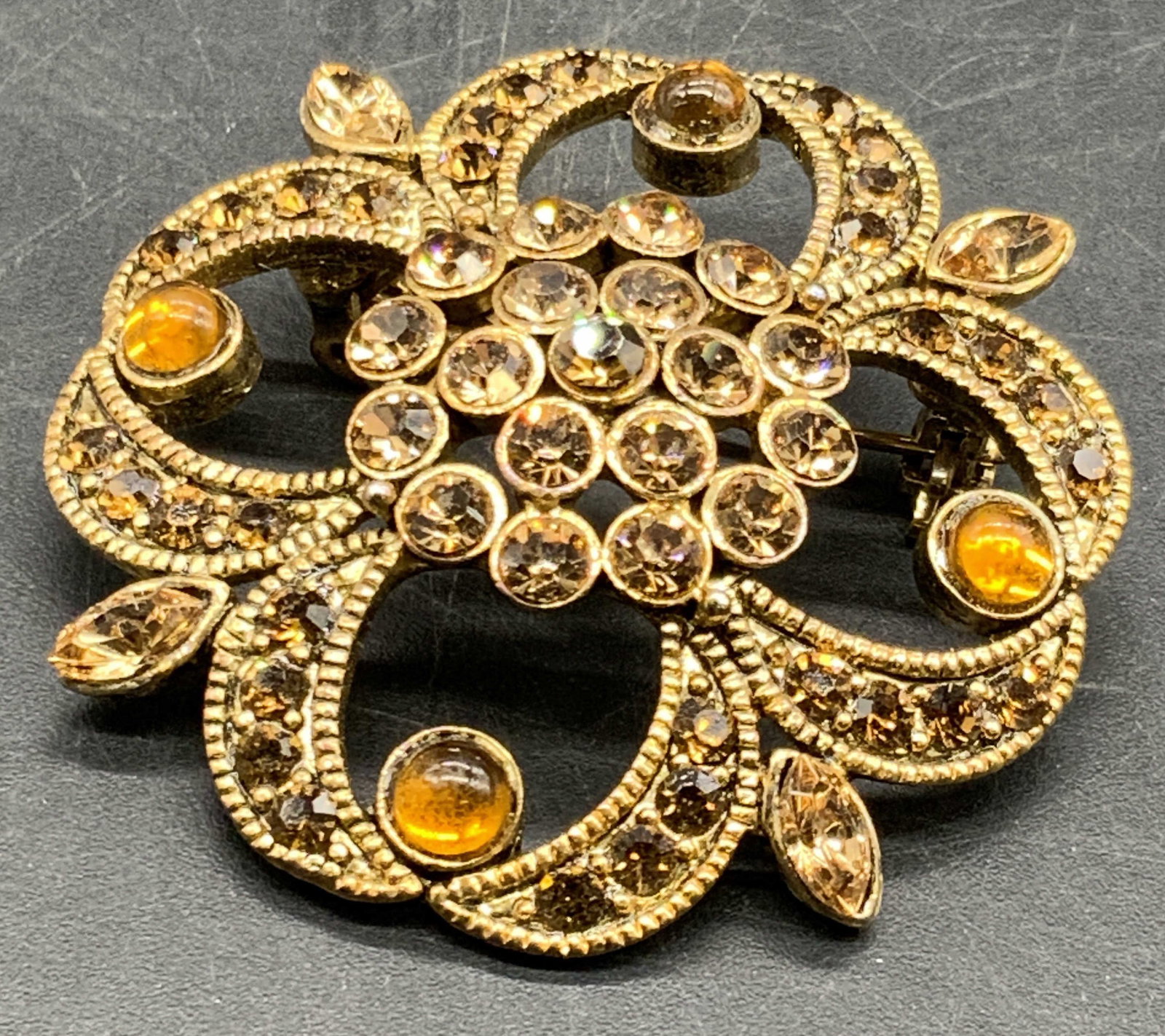 Vintage Bejeweled Crystal Abstract Form Brooch: A vintage gold tone metal abstract form brooch decorated with amber hued crystals and polished amber hued cabochons. Metal pin closure to reverse. Measures approx. 1.75L x 1.75W in. Some wear consiste