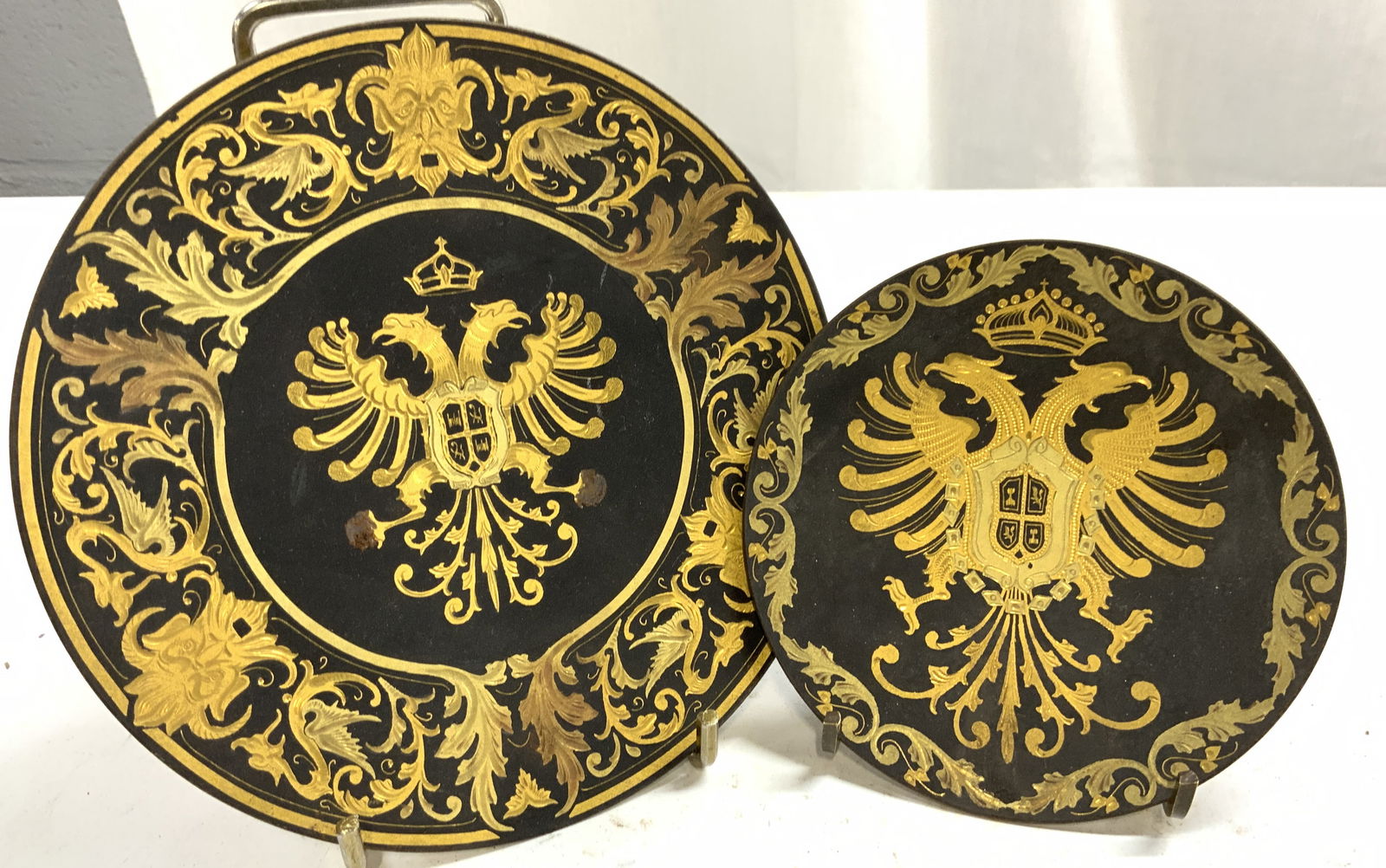 Lot 2 Spanish Damascene Plates: Lot includes 2 plates. Each Spanish Damascene. Plates are each black with gold toned gilt metal scroll, coat of arms, leaf & crown detail. Each plate has 3 feet. Largest plate measures approximately 6