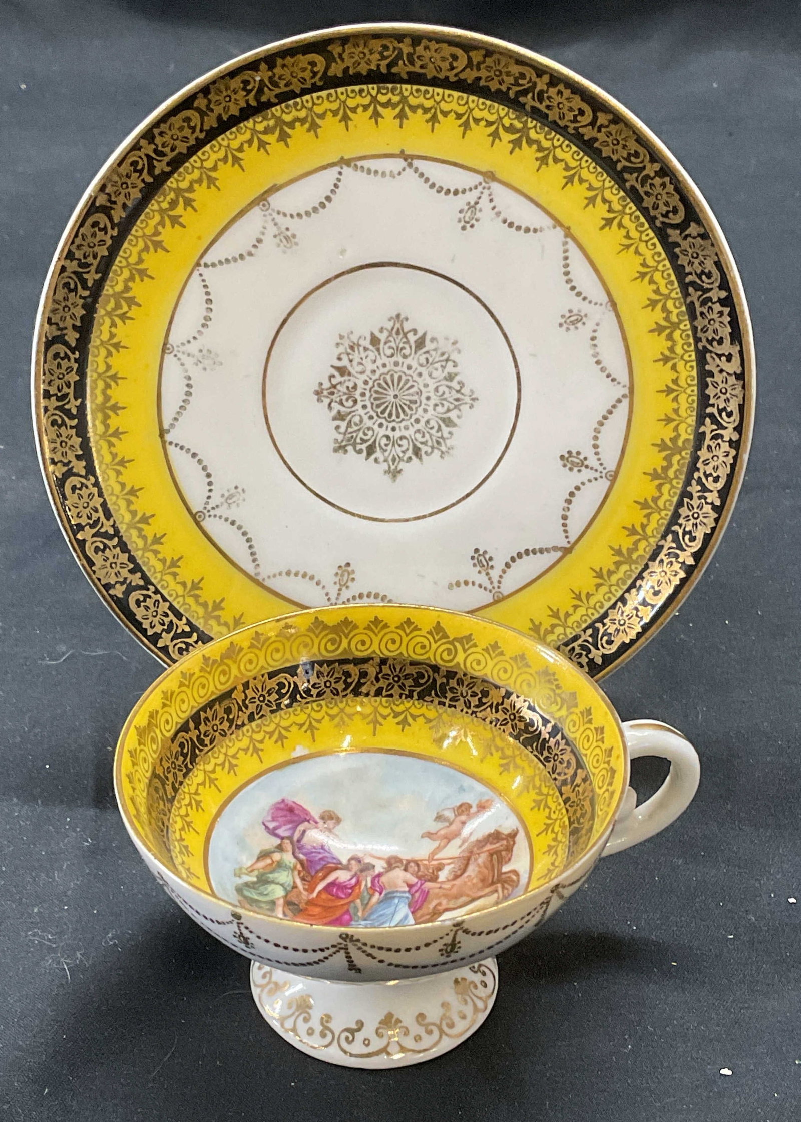 Signed Gilt Porcelain Demitasse & Saucer: Signed porcelain demitasse and saucer. Yellow and black with Grecian motif to center of cup. Gilt decoration. Hand painted. Hallmarked to underside of cup. Cup measures approx. 2.75 x 1.75 inches, sau