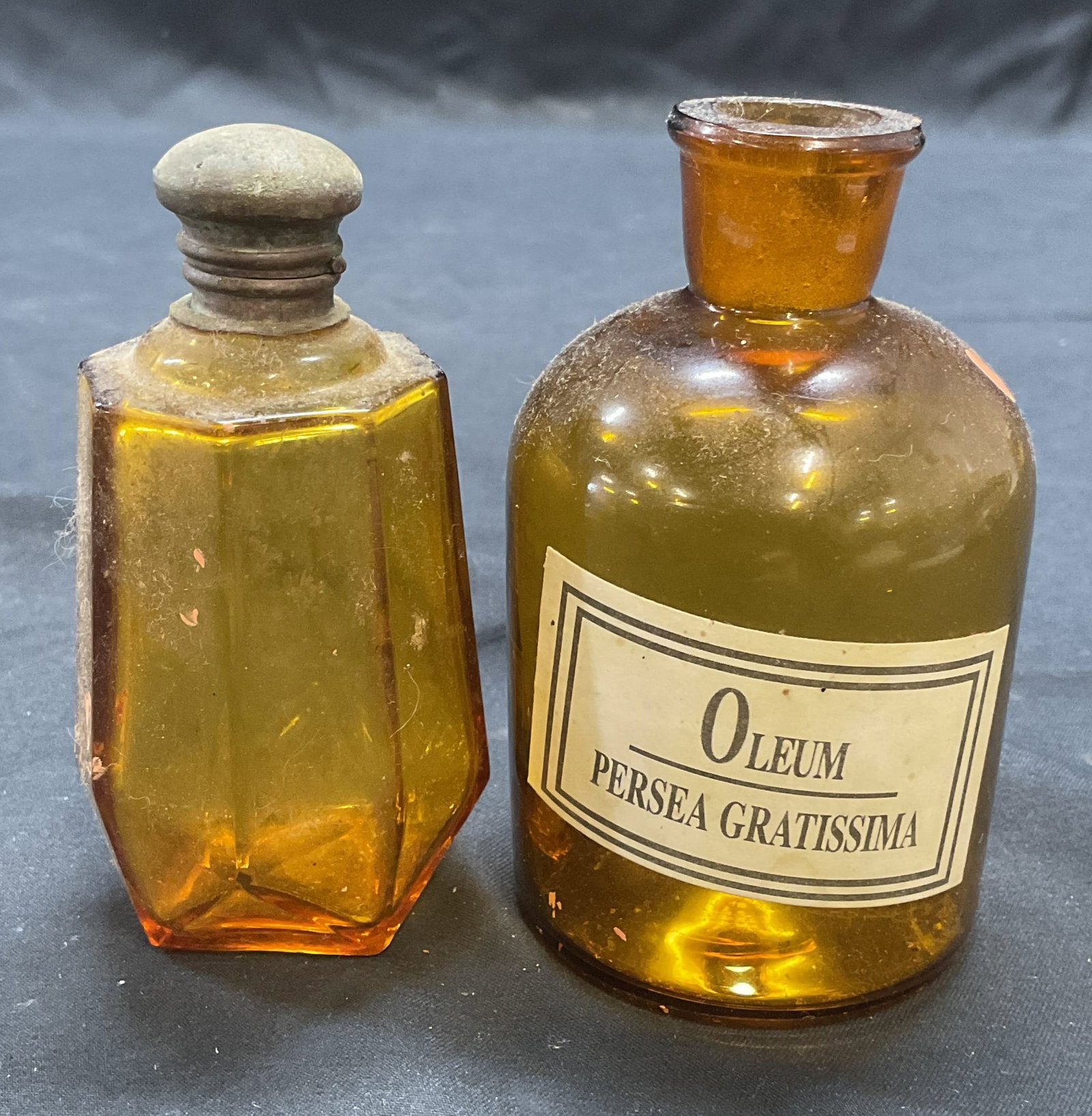 Lot 2 Vintage Avon Amber Glass Bottle +: Lot of 2 vintage amber glass bottles. 1 bottle marked Avon on underside. 1 bottle with label that reads Oleum Persea Gratissima. Larger bottle measures approx. 2 x 3.75 inches. Both pieces have surfac