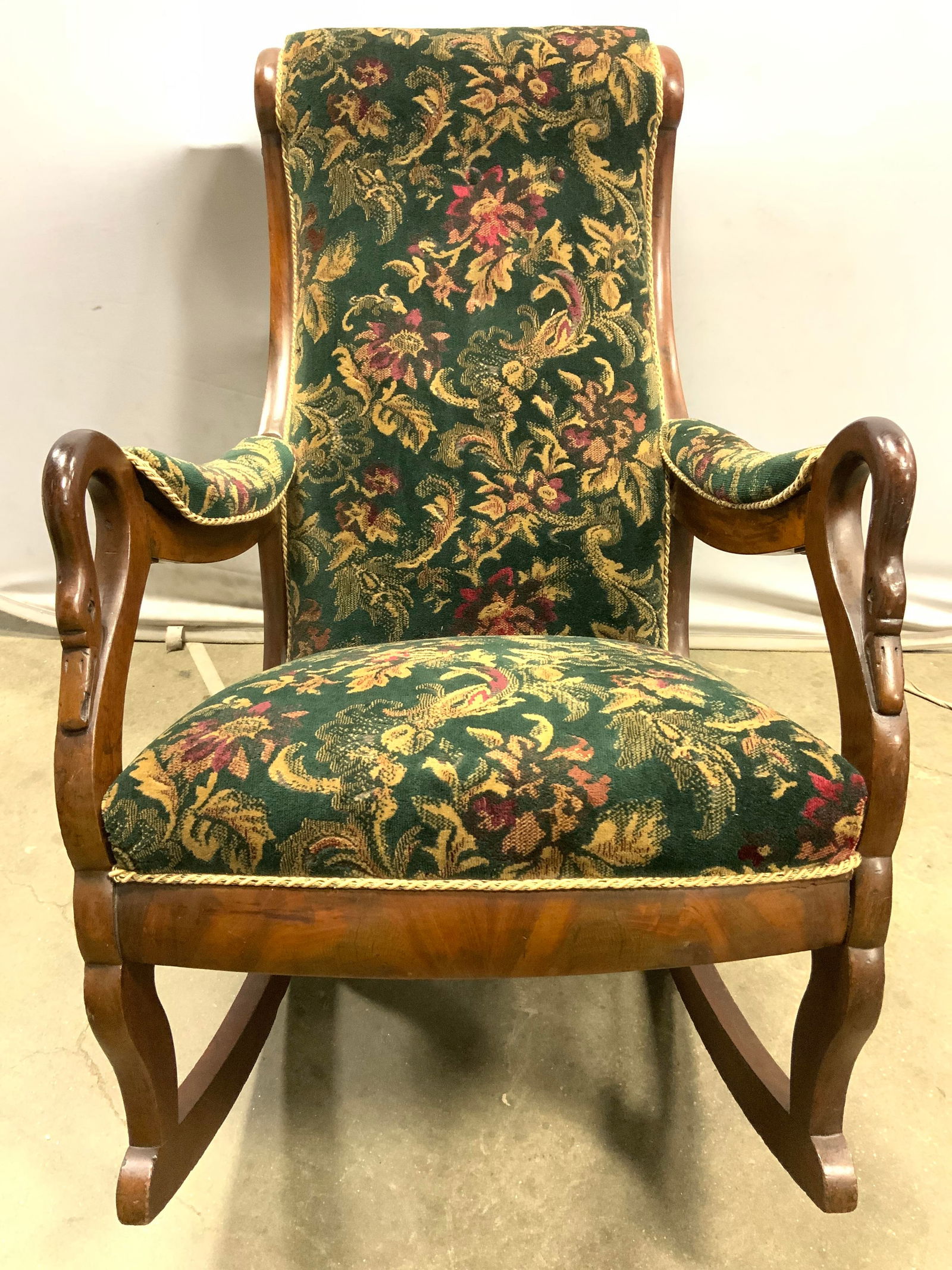 Antq Edwardian Swan Head Uph Wooden Rocking Chair: Rocking chair is antique and wooden. Piece has carved swan head detail to ends of armrests. Chair has multi toned floral and leaf upholstery with gold toned corded edges. Measures approx 40 inches dee