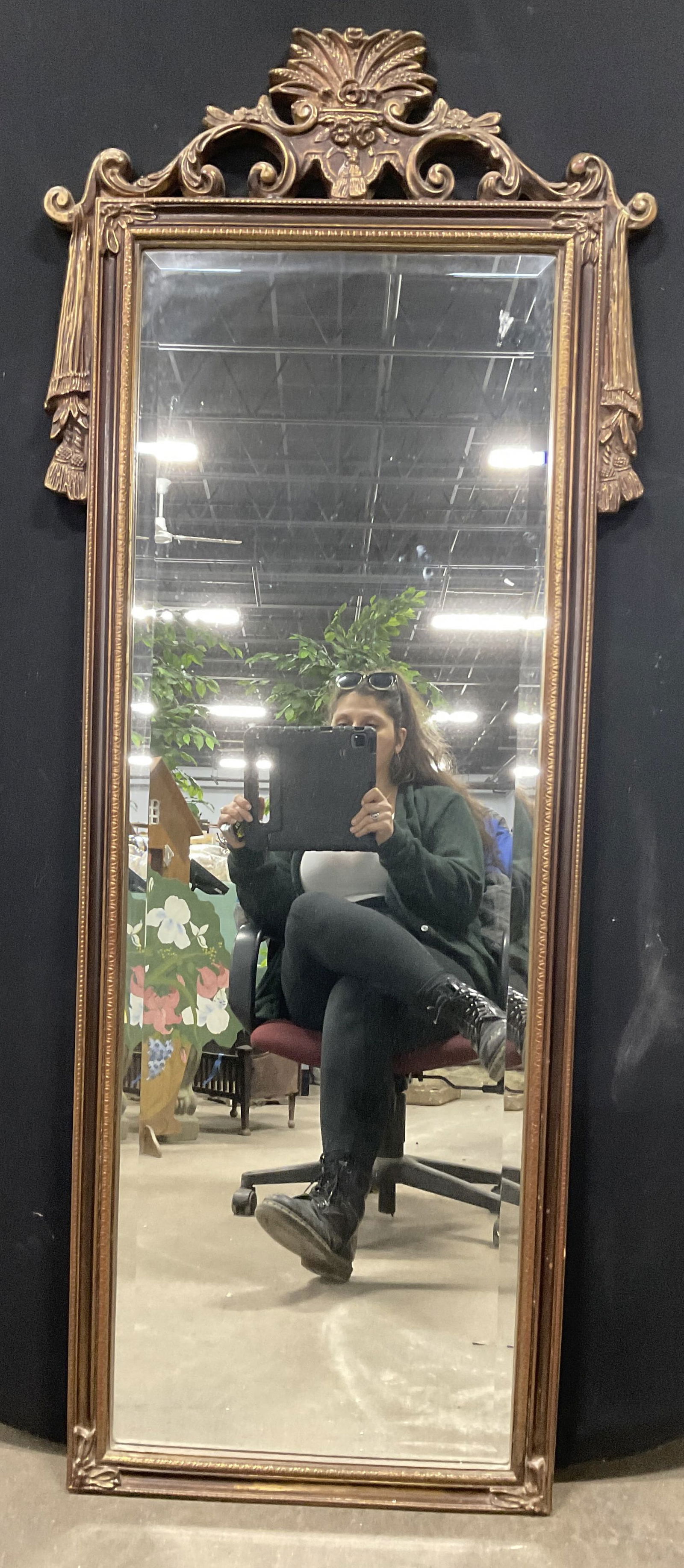 Gilt Wood Floral Motif Mirror: A gilt wood mirror with floral and scrolled woodwork designs. Red hues throughout the gilt. Measures approx. 61L x 25W in. Some surface wear consistent with age, use, and exposure. Discoloration prese