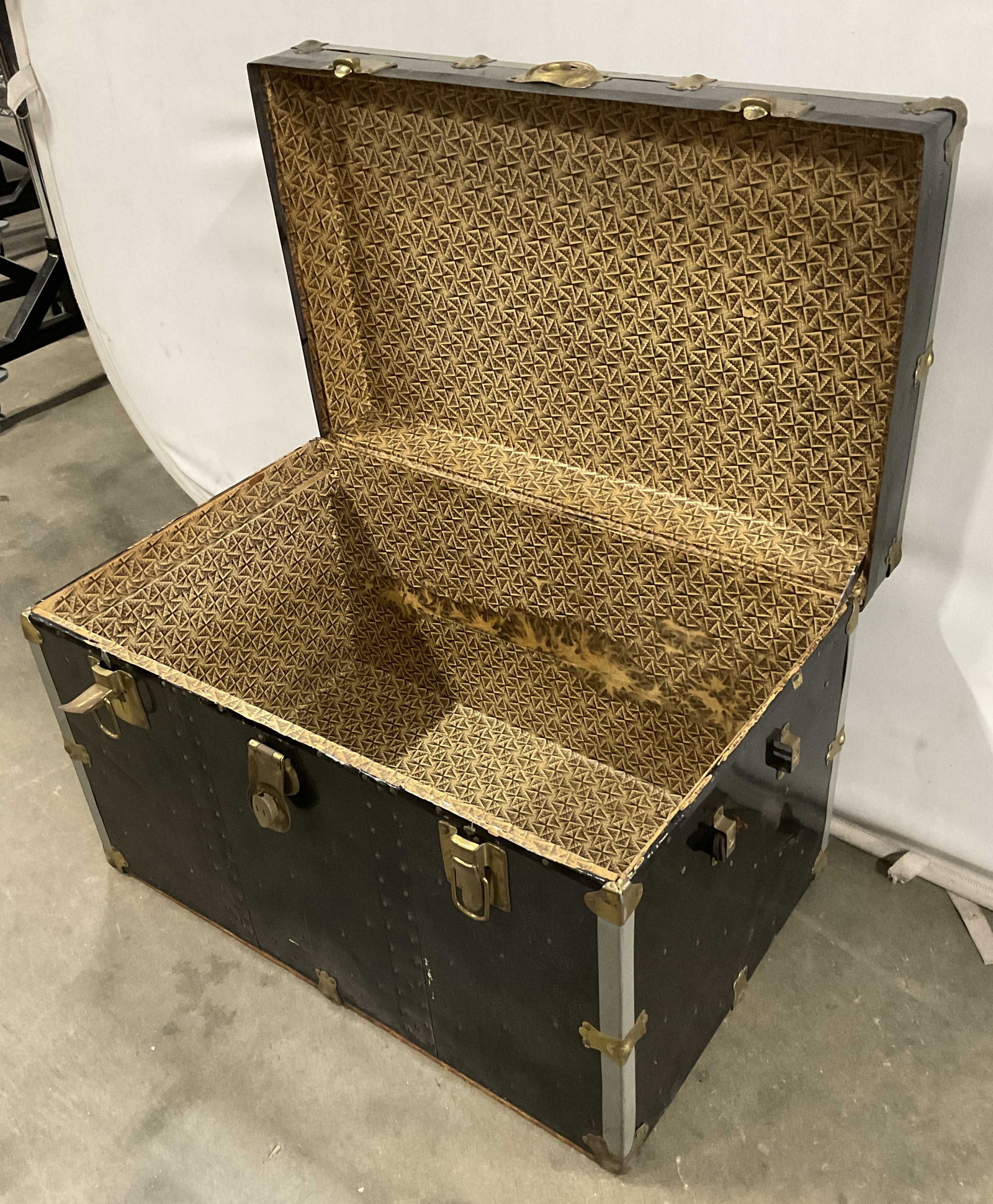 Vintage Black Wooden Travel Trunk: A vintage black wooden travel trunk. Deep brass hued metal hardware. Handles on either side, one handle needs repair / replacement. Lid opens with hinge hardware. Measures approx. 19H x 30W x 19D in.