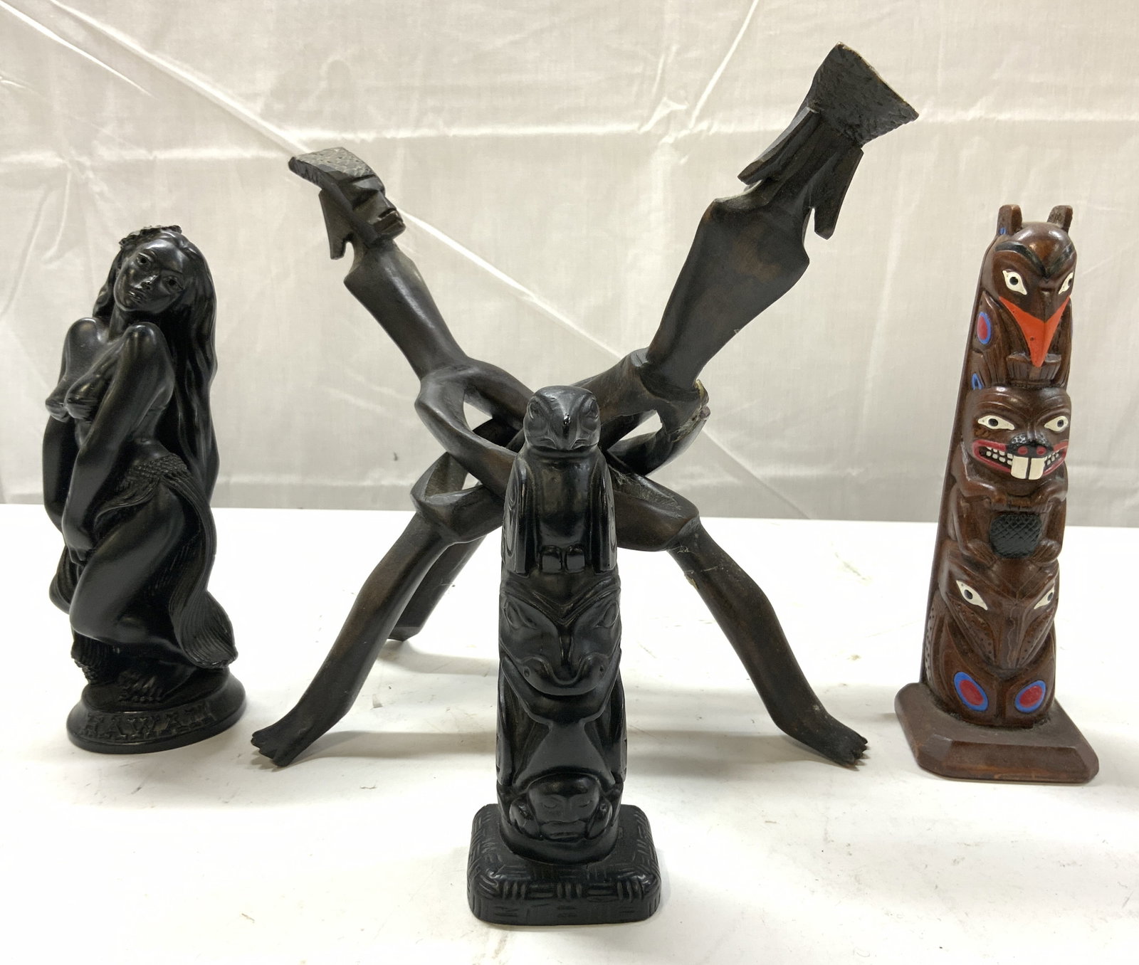 Lot 4 Interlocking Sculp & Other Tribal Carvings: Lot includes 1 dark chocolate toned interlocking figure sculpture, 1 female figure, base reads HAWAII, 1 Brown toned wooden tribal totem, 1 black toned resin tribal totem. Stand measures 9.5 inches ta