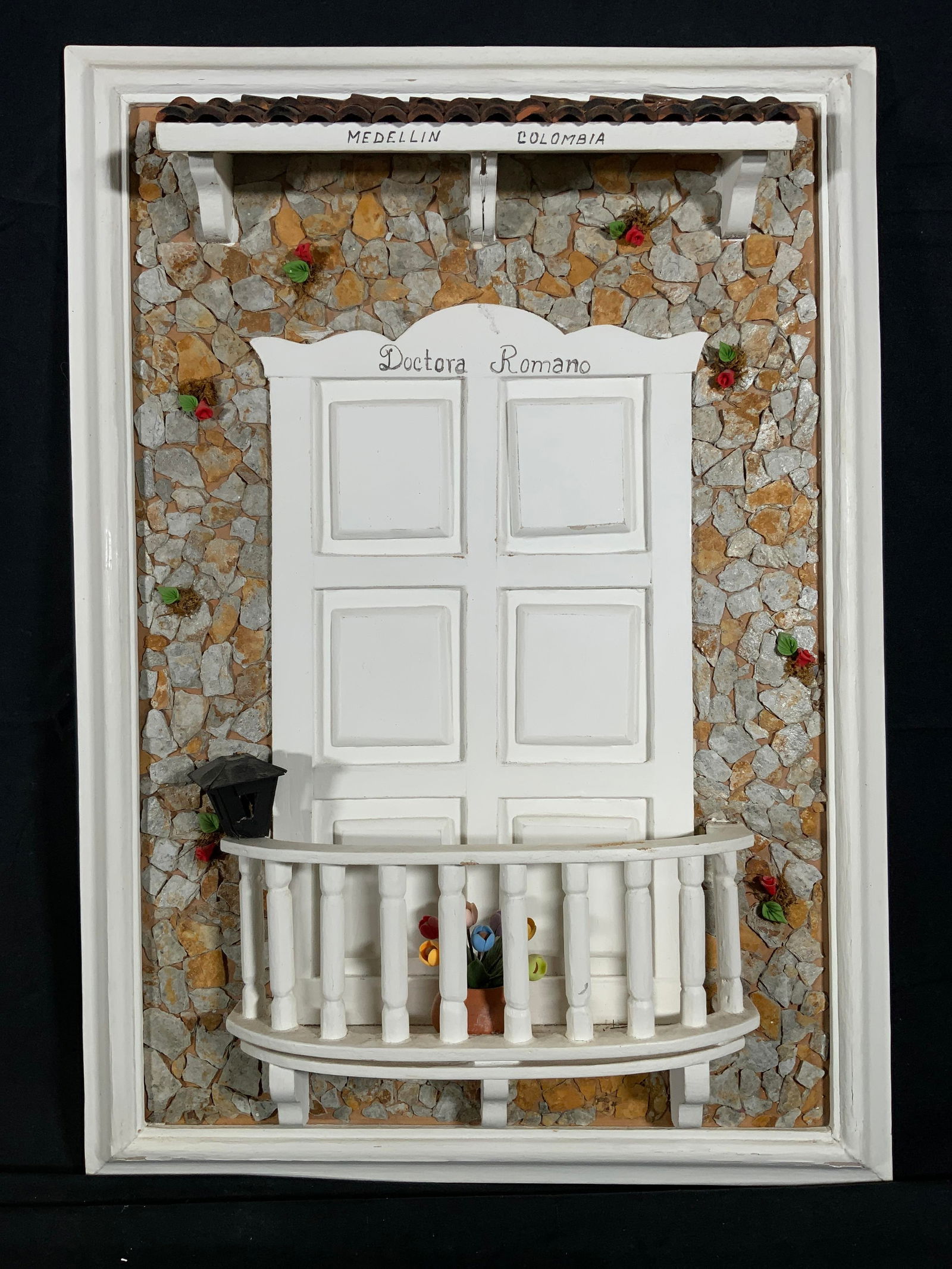 Wooden Colombian Balcony Door Wall Hanging: Mixed media wall hanging relief. Subject depicts a white balcony door and small terrace of a stone building with a pot of flowers and a lantern sitting on the edge. Inscriptions read Mendellin Colombi