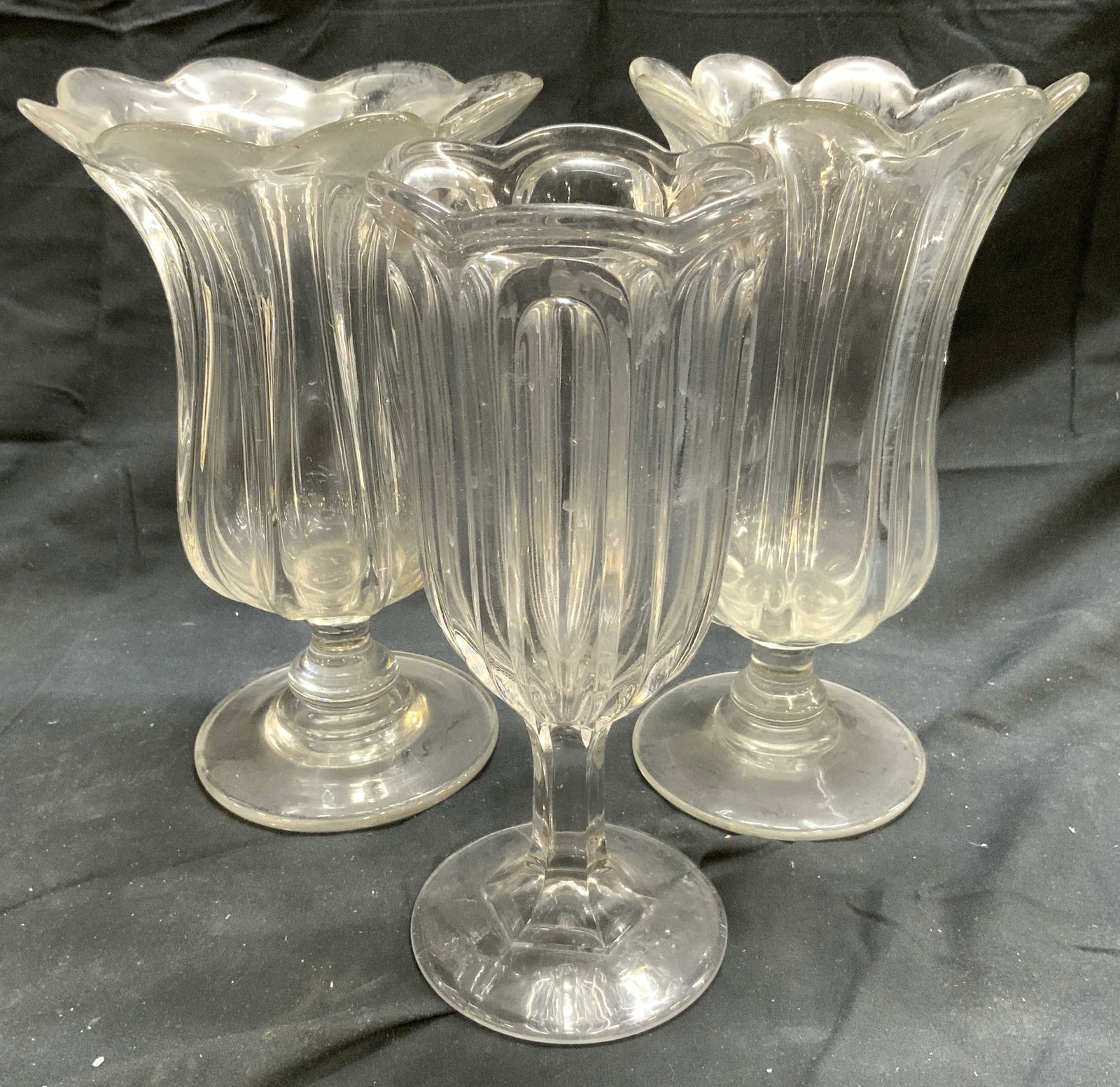 Lot 3 Pressed Glass Celery Vases Vessels: Lot includes 3 celery vase. Each piece is pressed glass. Vases each have stems with circular form bases. Each piece has floral form rim which lean outward. Largest vessel measures approximately 9 inch