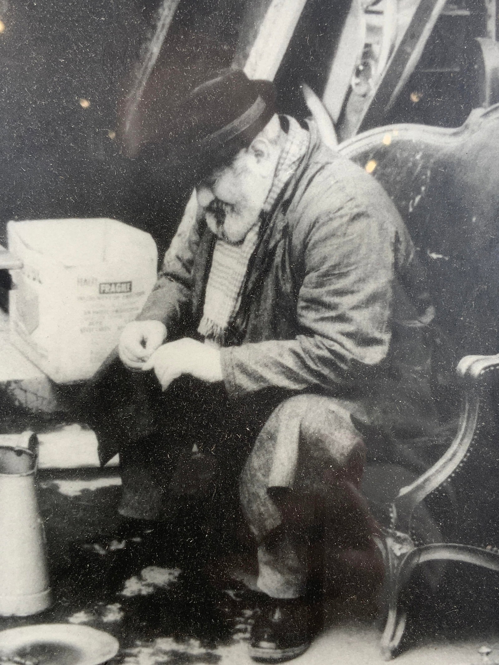 Framed Black & White Photograph Man Sitting: Black and white photograph. Subject depicts a man with a hat and scarf sitting on a large chair looking at his fingers. Framed in a gilt wooden frame. Frame measures approx. 11 W x 13 H in. Window mea