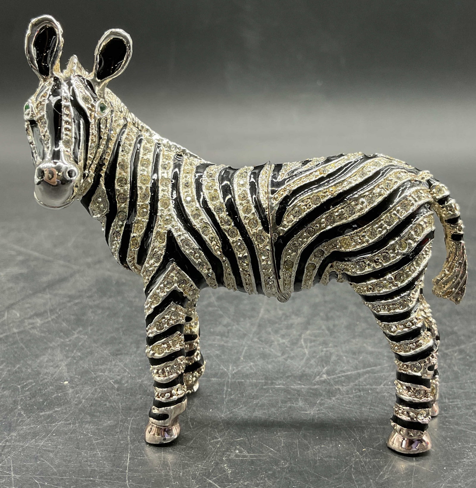 Bejeweled Enameled Metal Zebra Form Trinket Box: A metal zebra form trinket box bejeweled with crystals and decorated with black enamelwork. Green faceted crystals for the zebras eyes. Opens with hinge hardware. Measures approx. 3.5H x 3W in. Some s