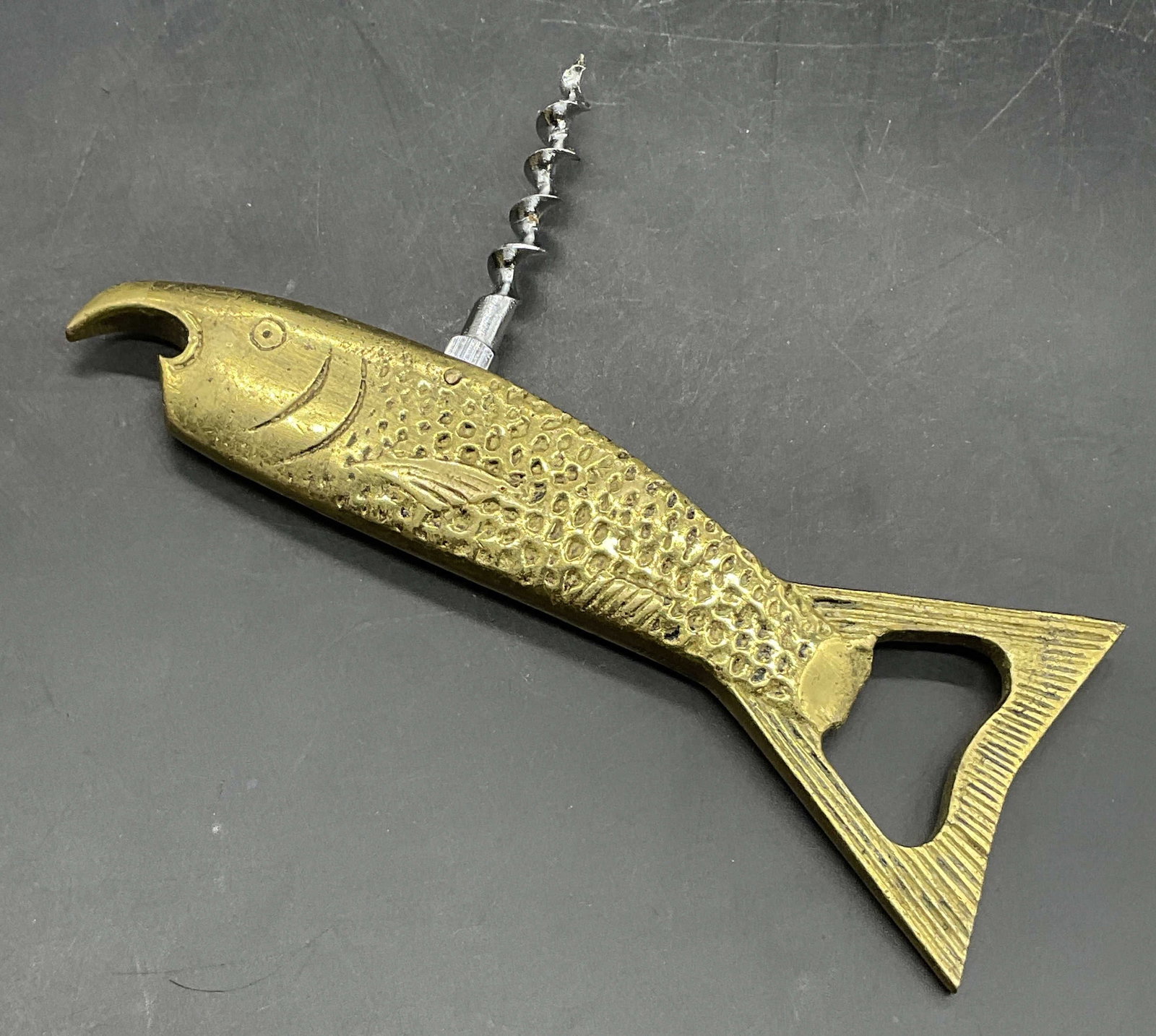 Brass Fish Bottle Opener: A brass fish form bottle opener. Silver tone metal corkscrew. Measures approx. 5.25W x 2L in. Surface wear consistent with age, use, and exposure. home decor, home accents, homewares, housewares, home