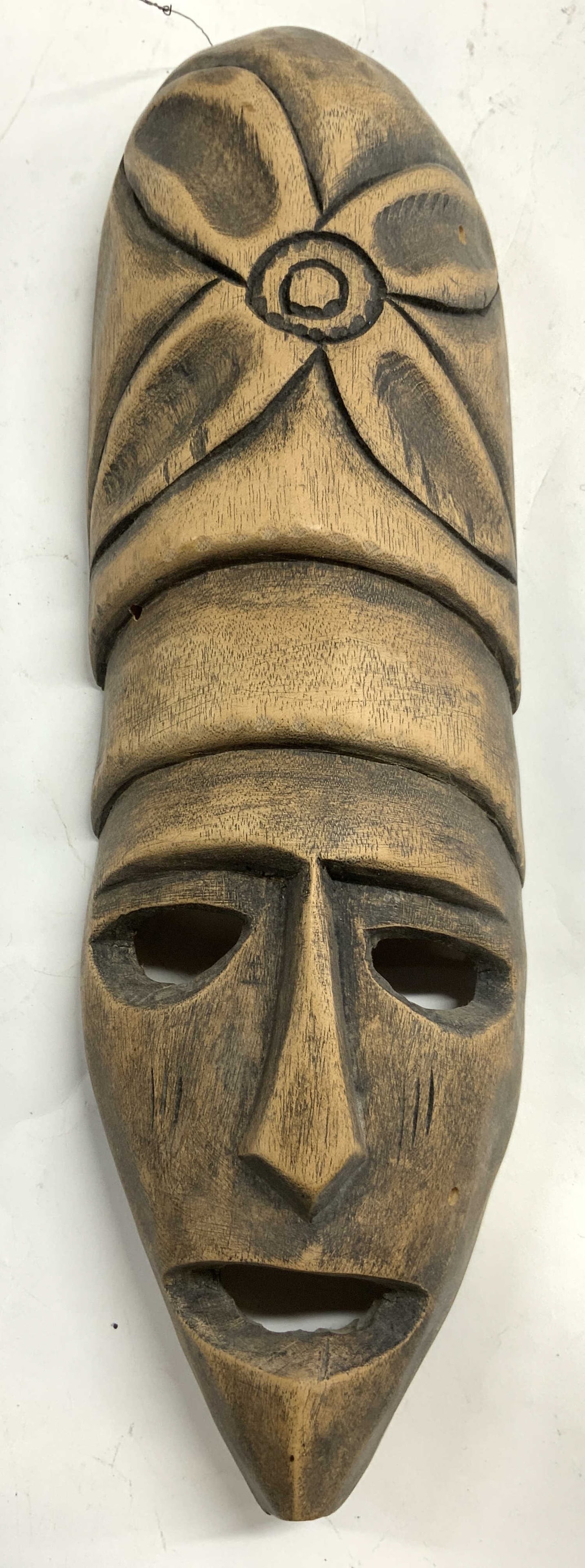 Vintage Carved Wooden Tribal Mask Wall Hanging: Wall Hanging tribal masks is tan toned and wooden. Piece has carved facial features, pierced eyes and mouth detail. Floral relief at top. Piece measures approximately 15cinches tall 4.5 inches wide. M