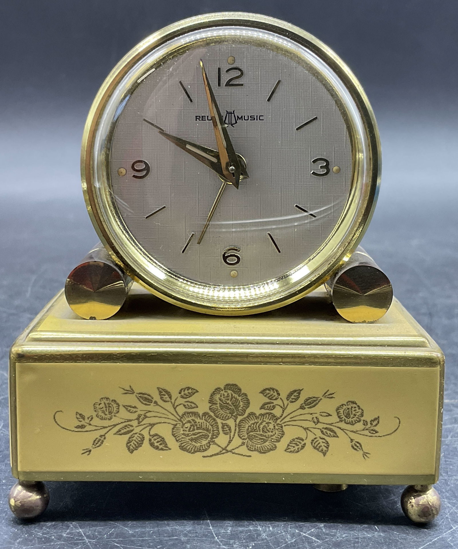 REUGE MUSIC Vntg Floral Motif Clock, Switzerland: A vintage metal clock with a floral motif to the obverse. Crafted from a brass tone metal with a yellow hued overlay. Face reads REUGE MUSIC. Underside reads MADE IN SWITZERLAND. Measures approx. 3.5H