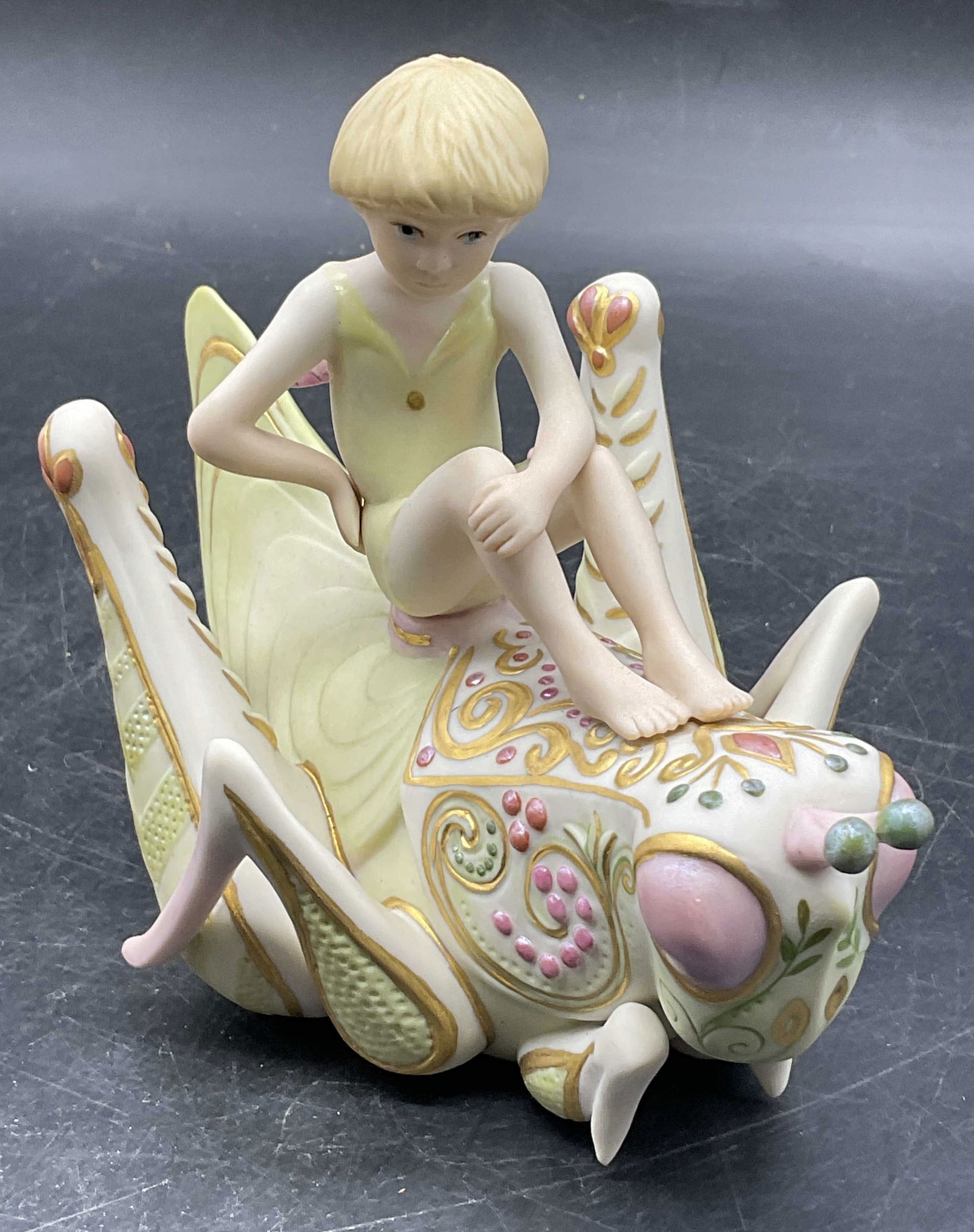 CYBIS USA Porcelain Child Atop Grasshopper Figural: A CYBIS USA Porcelain Fairy Child Atop Grasshopper Figural. Underside signed CYBIS, side reads CYBIS USA. Measures approx. 5H x 5W in. Some surface wear consistent with age, use, and exposure. home de
