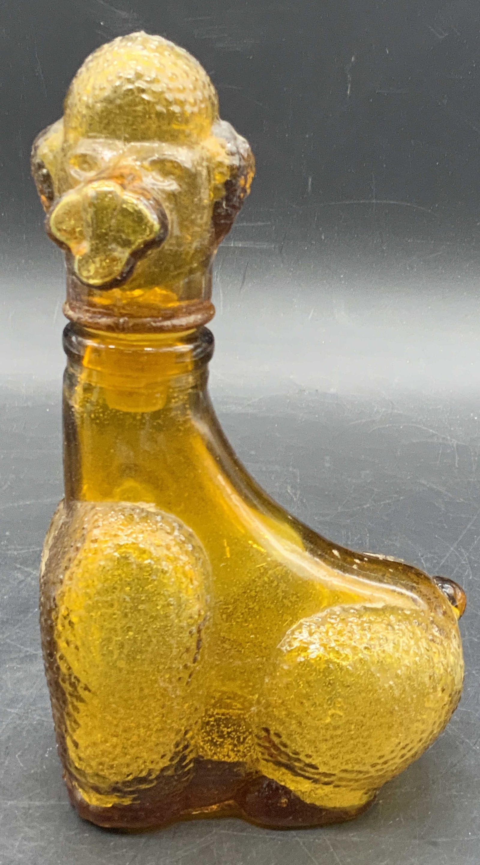 Vintage Amber Glass Poodle Dog Lidded Bottle: A vintage amber glass lidded bottle crafted to resemble a poodle. The dog measures approx. 7H x 3.5W in. Some surface wear consistent with age, use, and exposure. Seam and bubbles to glass present. Co