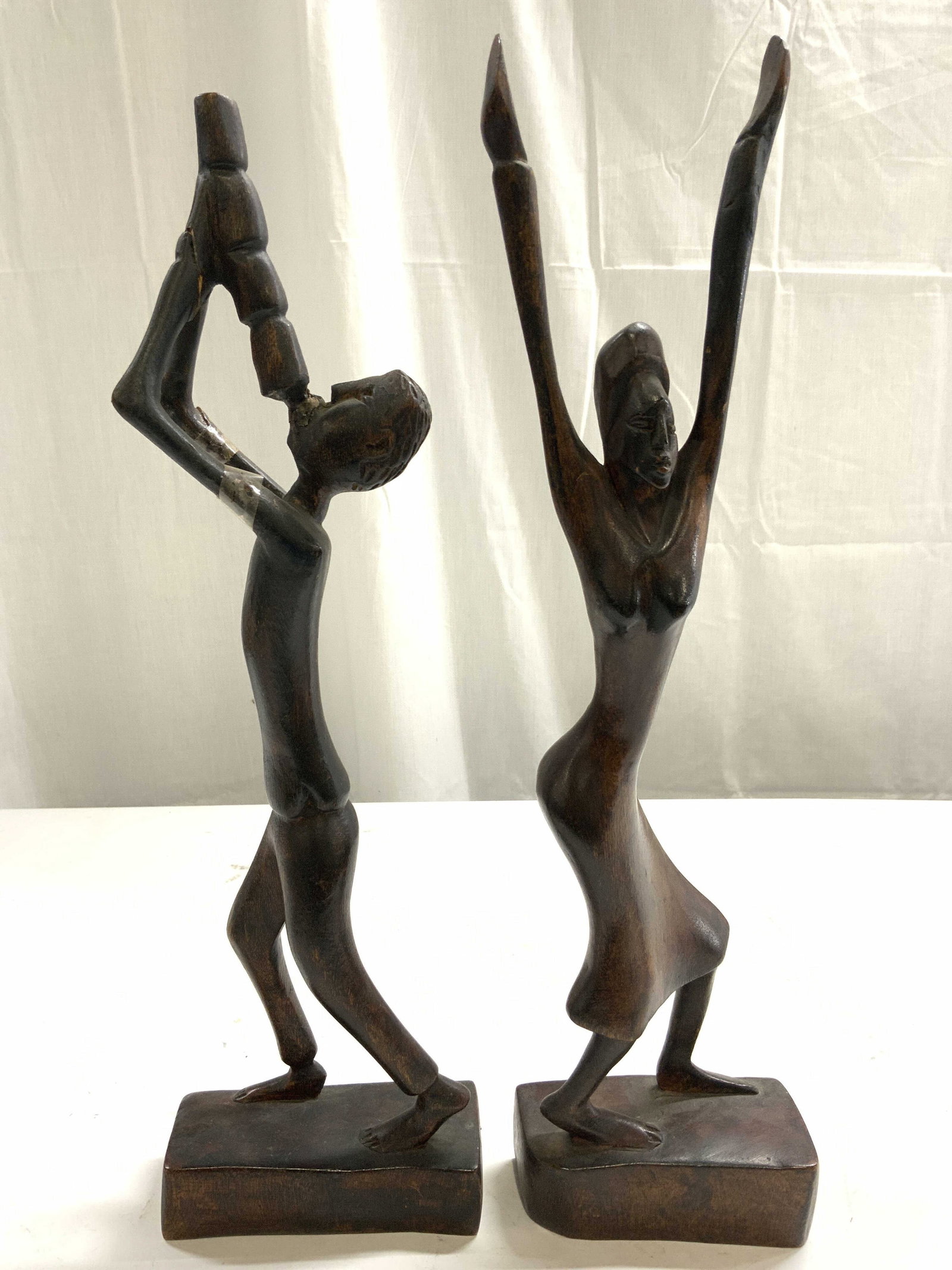 Lot 2 Wooden Male & Female Sculptures: Lot includes 1 female figure with arms stretched over head & 1 male figure playing an instrument. Each piece is brown toned and wooden. Female figure measures approximately 16 inches tall 4.5 inches w