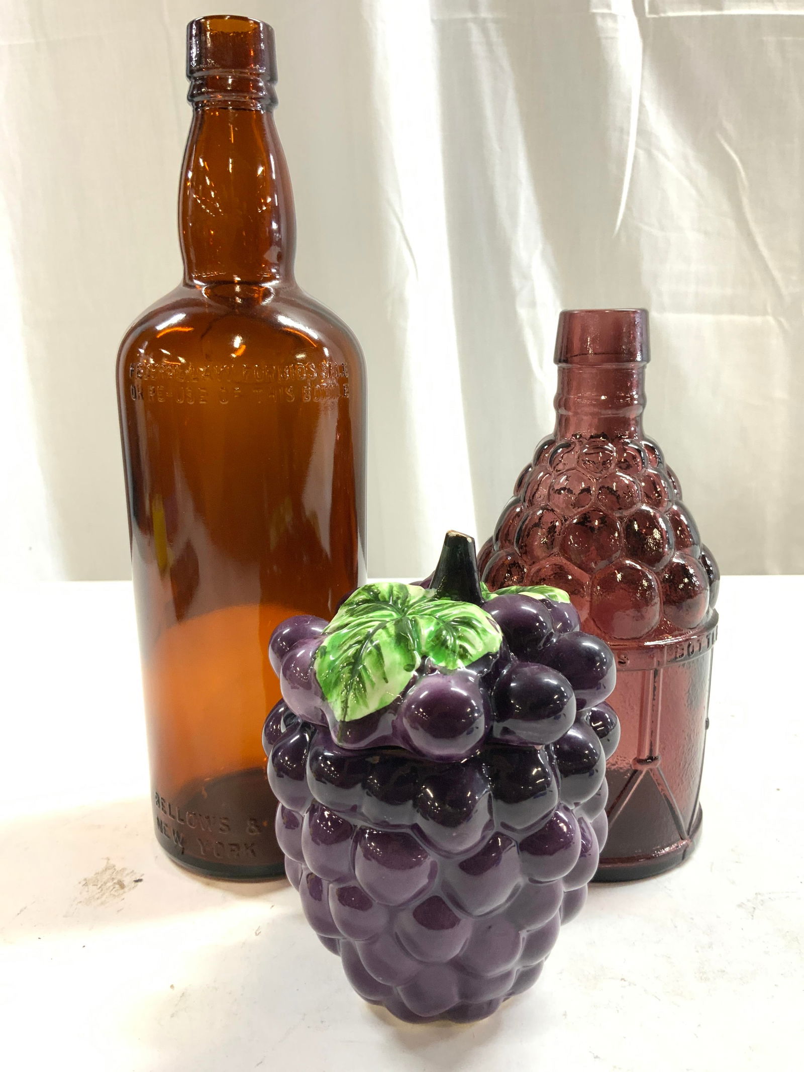 Lot 3 Art Glass Bottles & Porc Grape Jar: Lot includes 1 amber toned bottle which reads BELLOWS & COMPANY NEW YORK CHICAGO, 1 Purple toned bottle which reads MCGIVERS AMERICAN ARMY BITTERS BOTTLE & 1 purple toned porcelain grape form jar. Lar