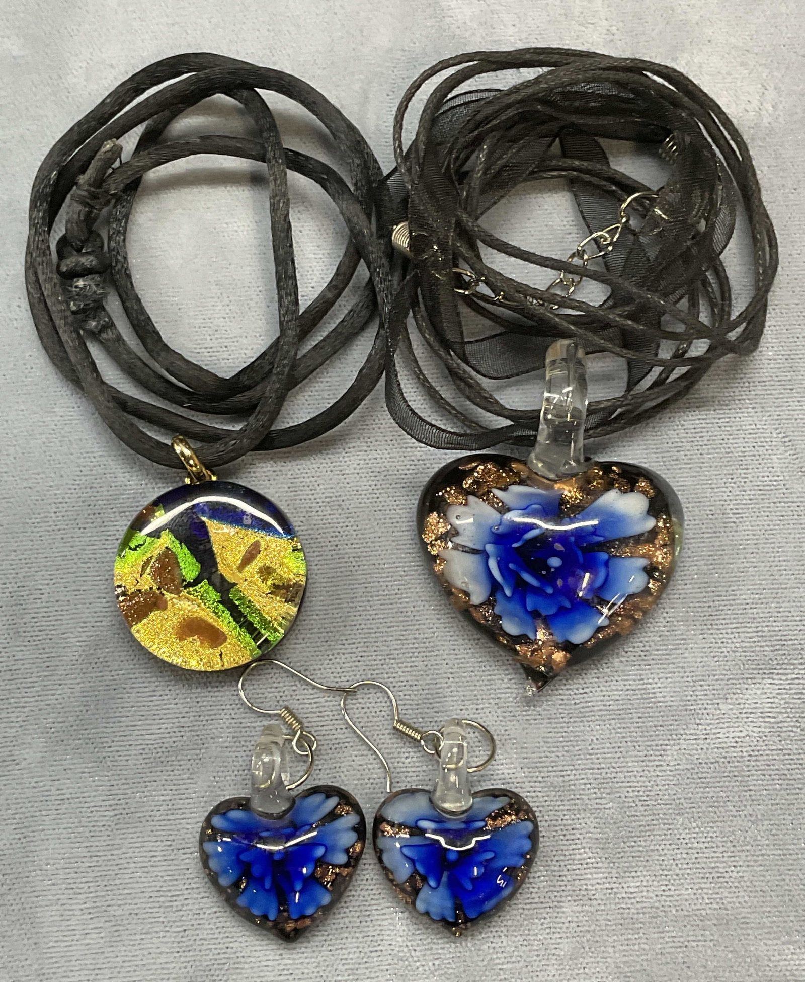Lot 3 Art Glass Heart Necklaces & Earrings: Lot of 3 jewelry pieces. Two art glass pendant necklaces on black cords. Heart and round shaped pendants. Drop length approx. 10 and 11 inches long. Art glass heart motif dangle earrings. Hook backing