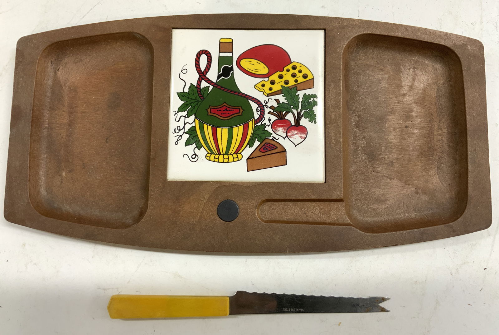 Lot 2 Wood & Porc Cheeseboard & Knife: Lot includes 1 wooden cheeseboard with porcelain tile in center. Tile has wine cheese & vegetable detail. Lot also includes 1 knife with Bakelite handle. Cheeseboard measures approximately 13 inches l