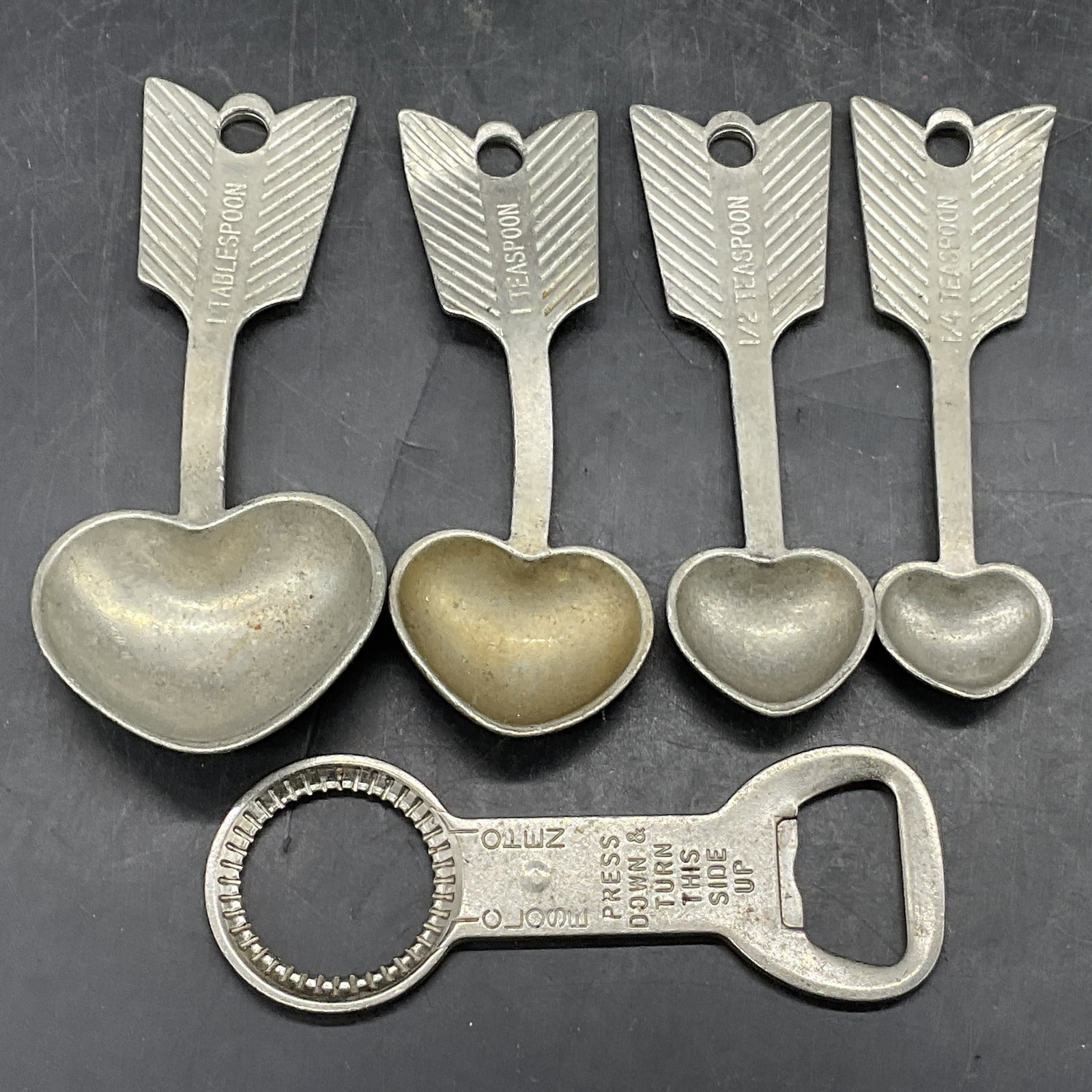 5 Vintage Metal Measuring Spoons & Bottle Opener: Four vintage metal measuring spoons and one metal bottle opener. The measuring spoons are tipped with heart and feather motifs. Largest measuring spoon measures approx. 4.5L x 2W in. The largest measu