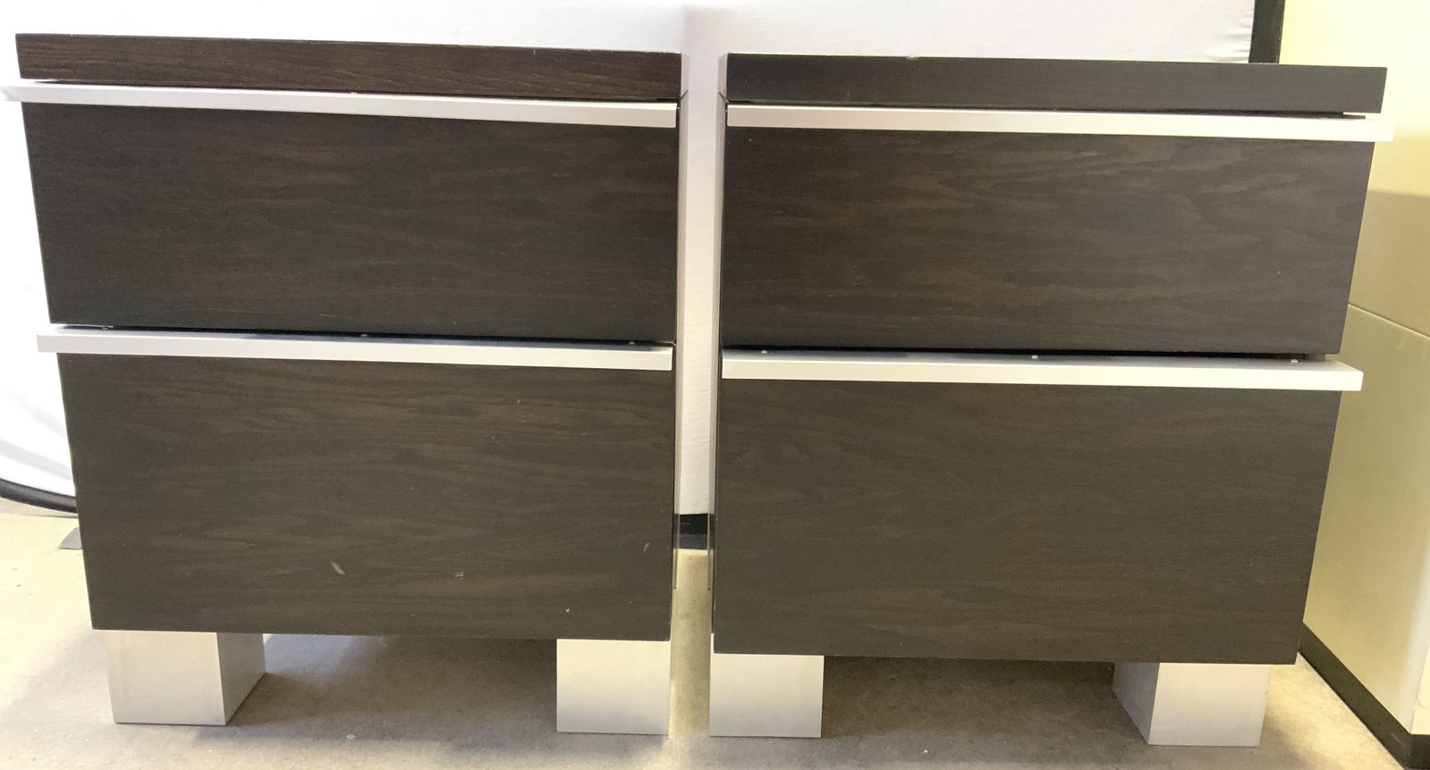 Pair Of Wooden Nightstands W 2 Drawers: Lot includes 2 nightstands. Each are brown toned & appears to be wooden. Nightstands each have silver toned metal handles. Nightstands have 2 drawers each. Each measures approximately 24 inches tall 2