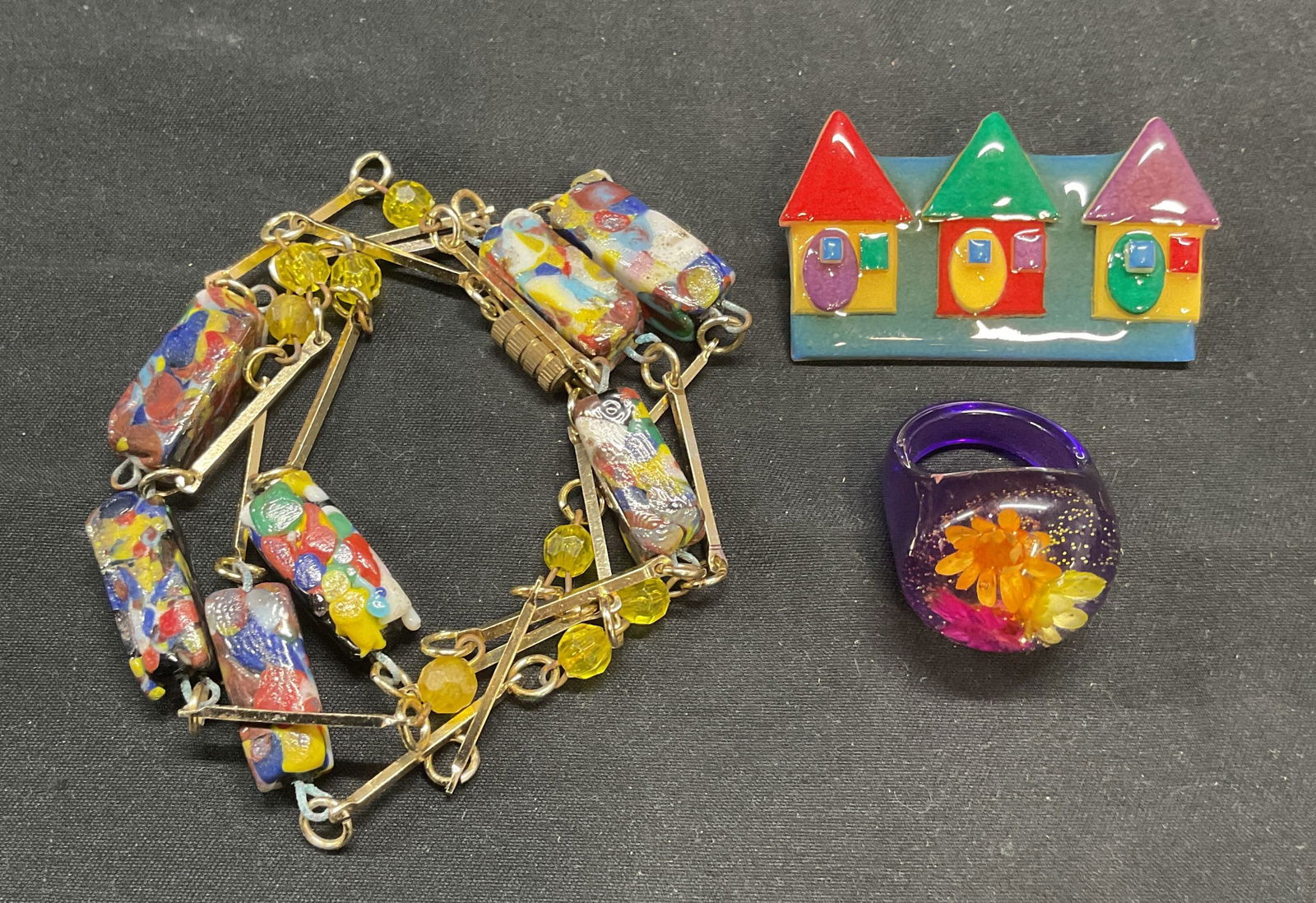 Lot 3 Art Glass Necklace, Flower Ring, +: Gold tone art glass beaded necklace. Drop length approx. 15 inches. Chain needs repair. Purple resin ring with flower decoration. Ring size 6.5. House Pins by Lucinda handmade house brooch. Measures a