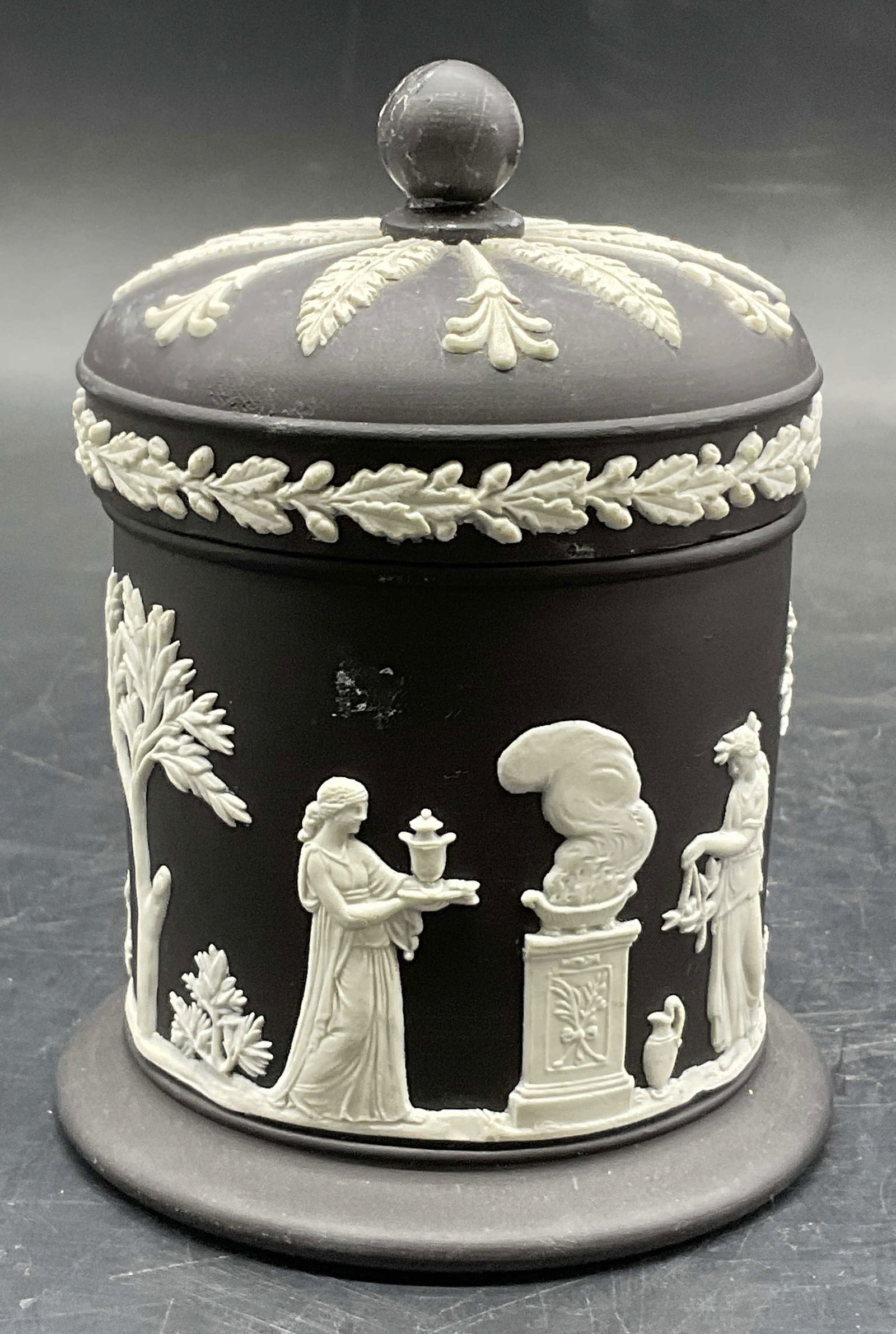 WEDGWOOD Sig Black Jasperware Lidded Vessel, Eng: A vintage black Jasperware lidded vessel. Raised white hued Greco Roman motifs throughout. Measures approx. 4.5H x 3.5W in. Measurement includes lid. Underside signed WEDGWOOD ENGLAND. Surface wear, c