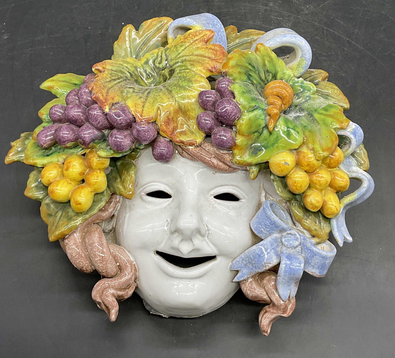 Vintage Signed Majolica Pottery Bacchus Face Mask: A vintage majolica pottery Bacchus face mask. Colorful ribbons and fruits to the face mask. Underside signed illegibly. Measures approx. 4D x 10.5W x 10L in. Pitting present. Drilled holes and wire to