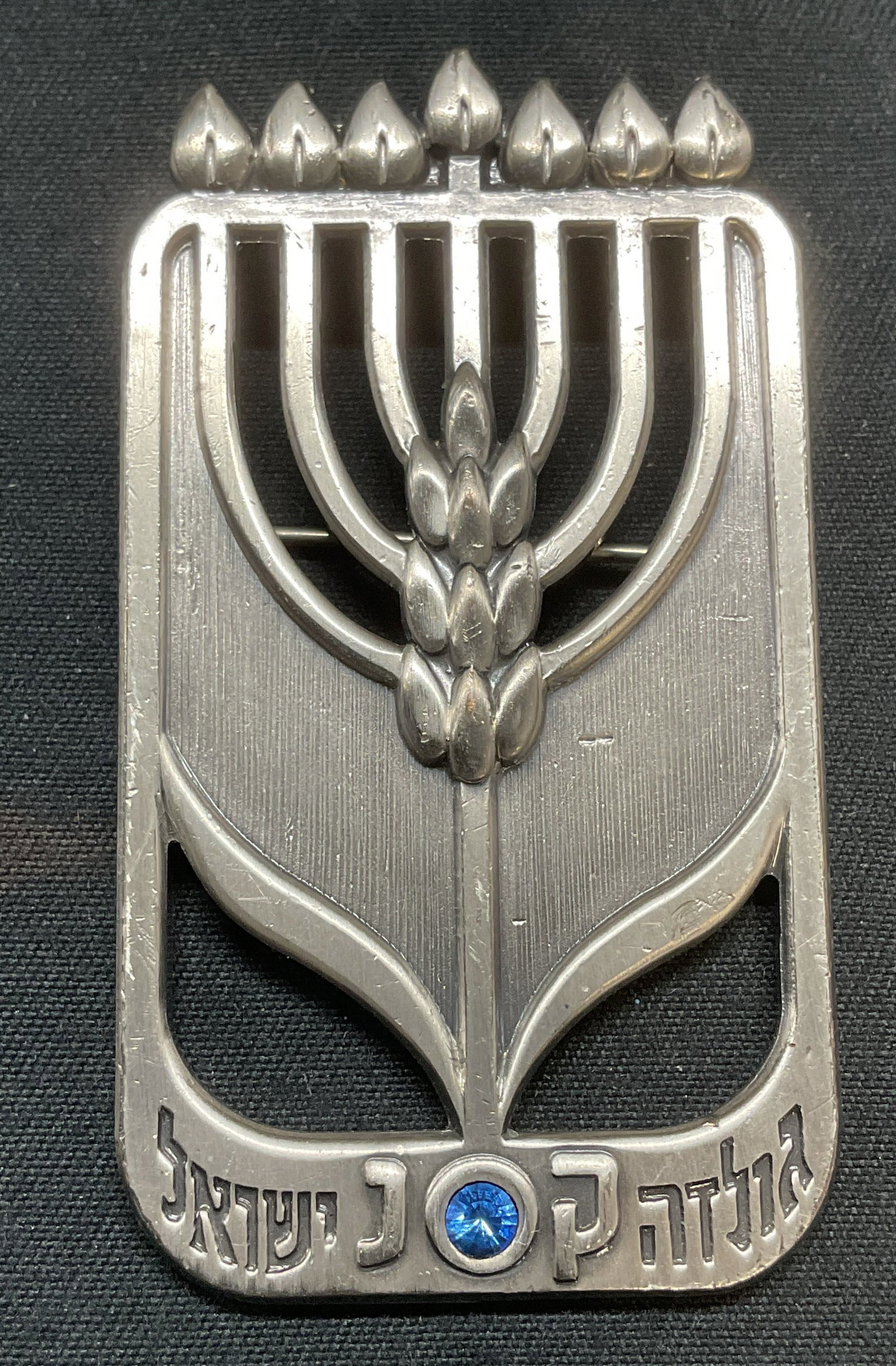 Avi Biran Judaica Menorah Silver Tn Brooch Israel: Avi Biran Judaica brooch. Silver tone menorah design with blue crystal. Signed on underside. Made in Israel. Hinge pin and bail on underside. Measures approx. 2.25 x 1.25 inches. Some surface wear inc