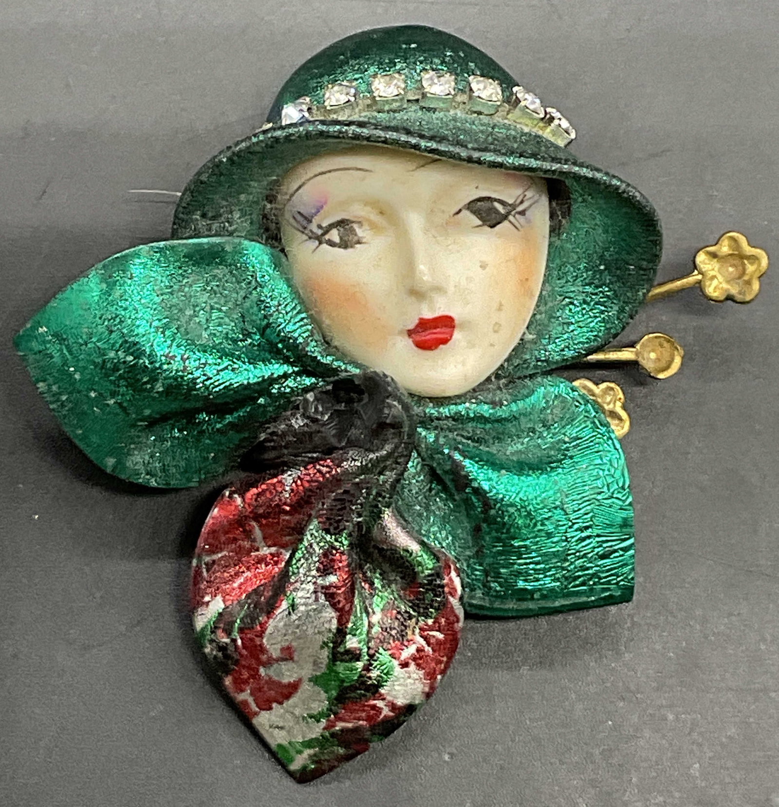 Vintage Lady Face Figural Brooch: A vintage lady face figural brooch. Crafted from metal and composite materials. Crystal accents to the womans hat and shimmery coloring to the rest of her outfit. Metal pin closure to reverse. Measure
