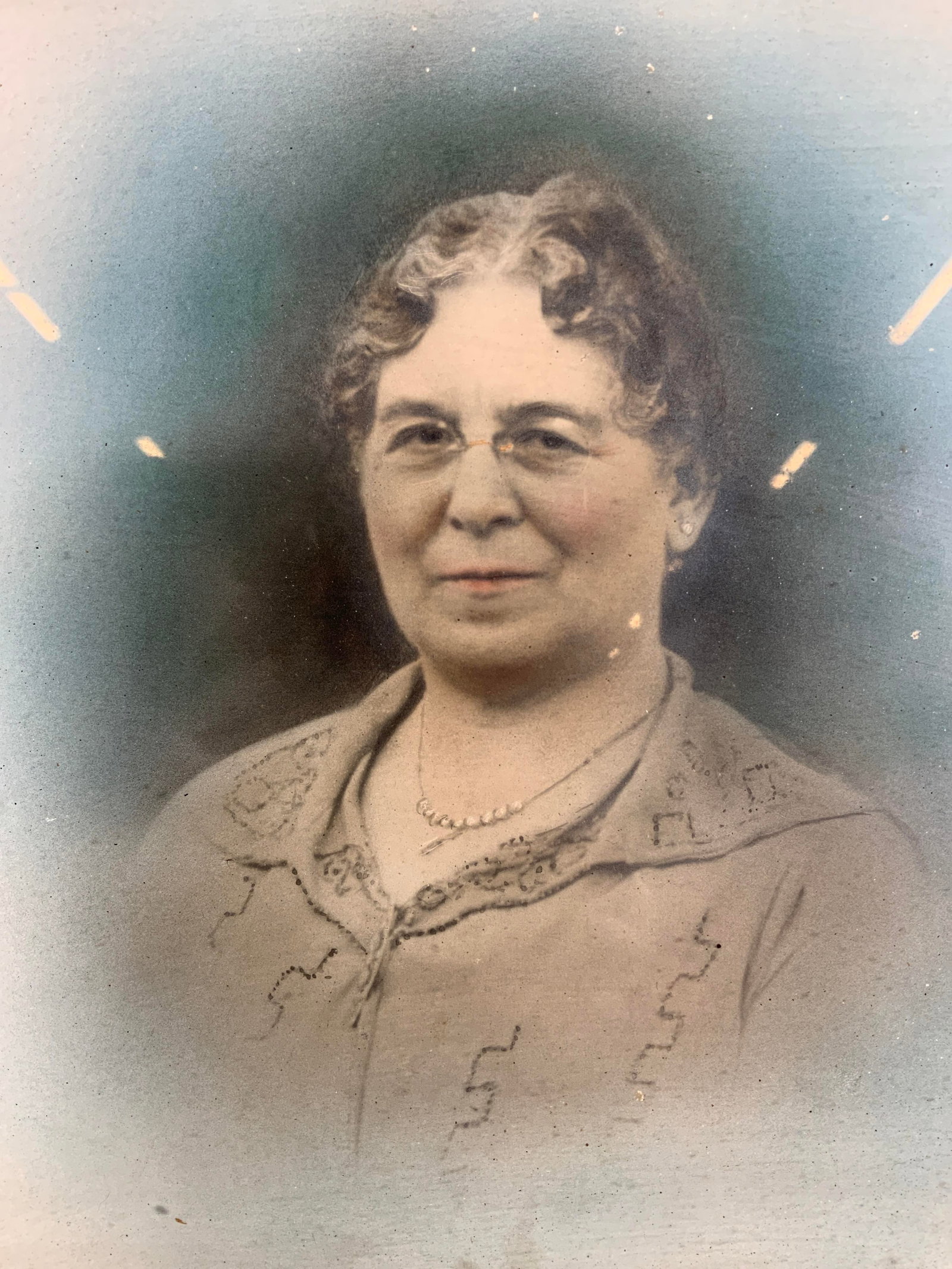 Framed Hand Colored Photograph of an Older Woman: Hand colored photograph. Subject depicts and older woman with glasses and a pearl necklace. Framed in a white wooden frame. Frame measures approx. 12.5 W x 15 H in. Window measures approx. 10.5 W x 12
