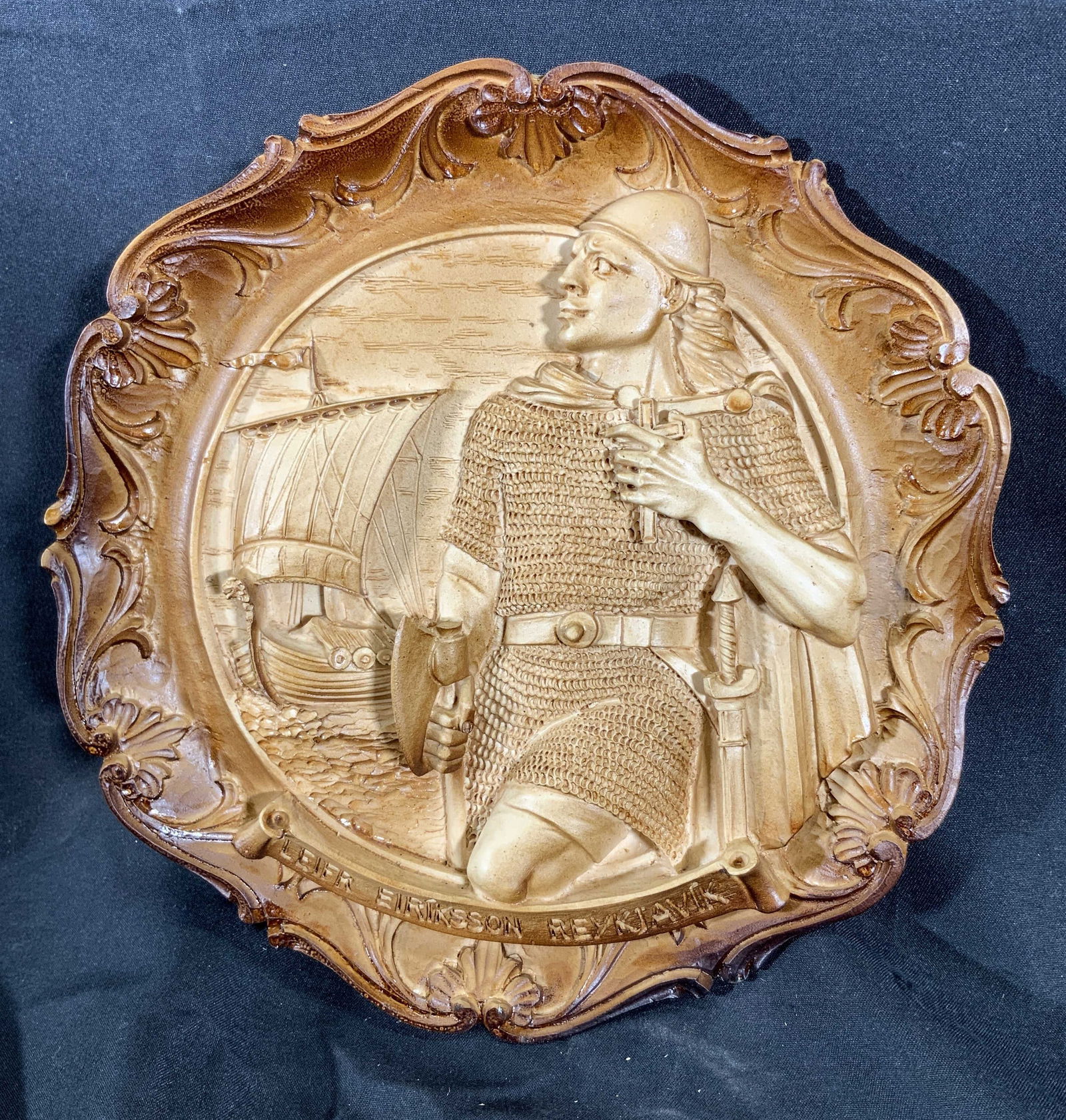 Leifr Eiriksson Reykjavik Viking Resin Plaque: Resinous wall plaque. Banner reads Leifr Eiriksson, Reykjavik. Subject depicts leifr Eiriksson the Viking posed with battle armor and an axe in front of his ship. Border of the plaque have scrolling m