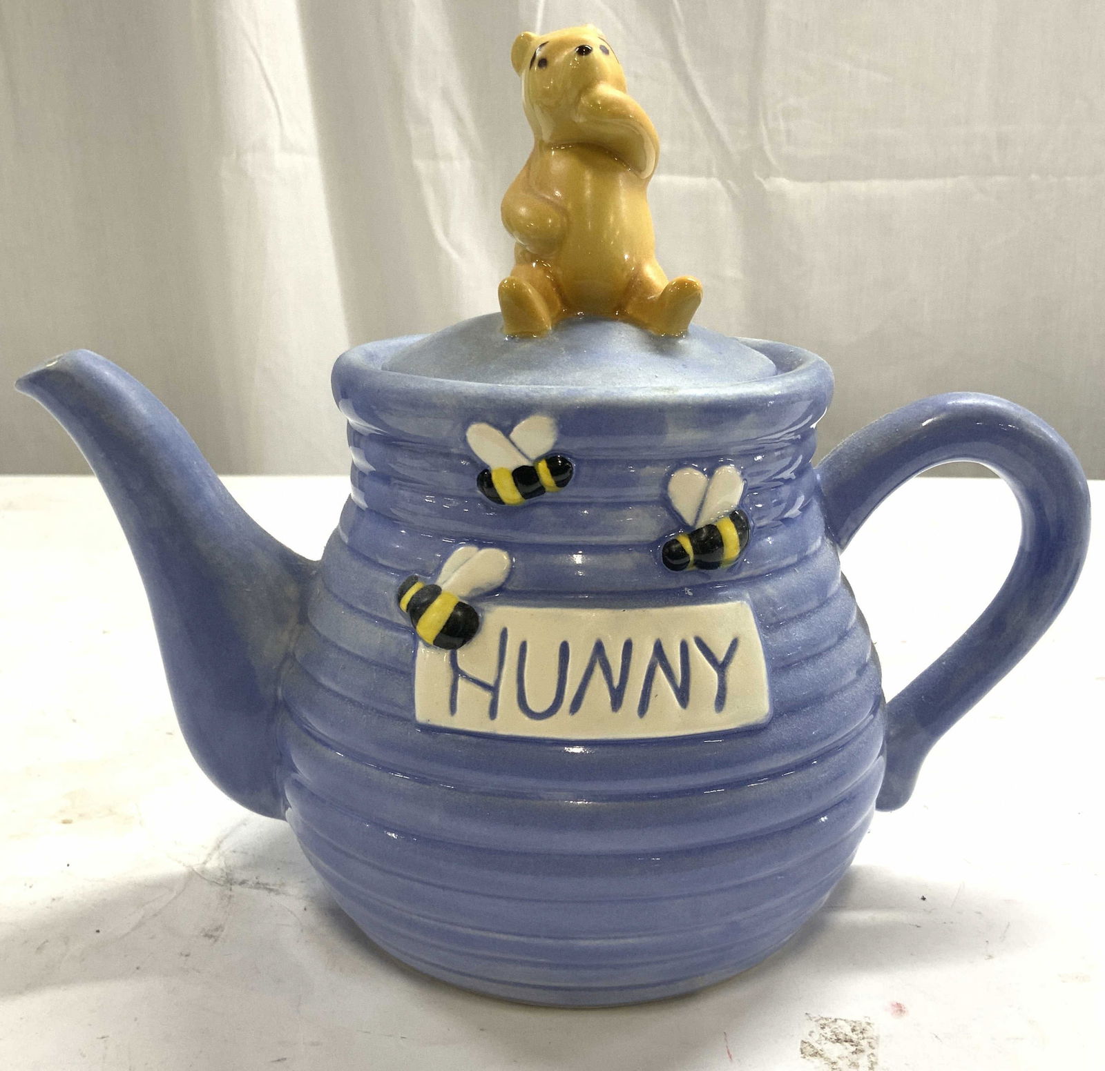DISNEY TREASURE CRAFT Winnie The Pooh HUNNY Jar: Underside reads DISNEY TREASURE CRAFT. Front side of teapot reads HUNNY. Teapot includes lid. Top of lid has Disney character Winnie The Pooh. Teapot is light blue toned and porcelain. Piece measures