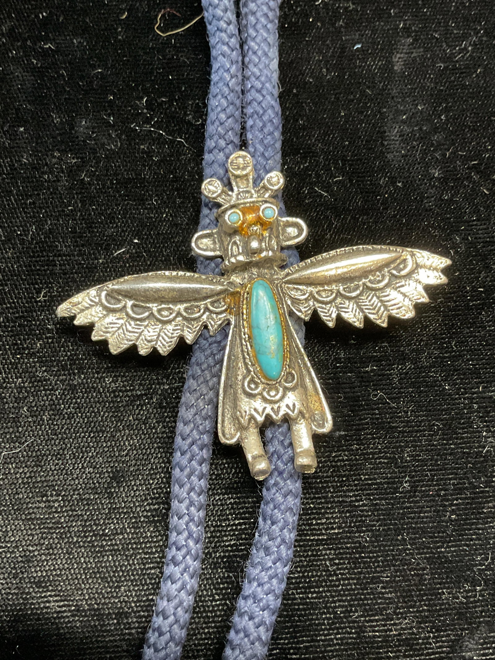 Silver Tn Tribal Figural Bolo Tie Necklace: Bolo tie necklace with silver tone tribal figural pendant and faux turquoise cabochon. Blue cord. Drop length approx. 16 inches. Pendant measures approx. 2 x 2.5 inches. Some surface wear including sc